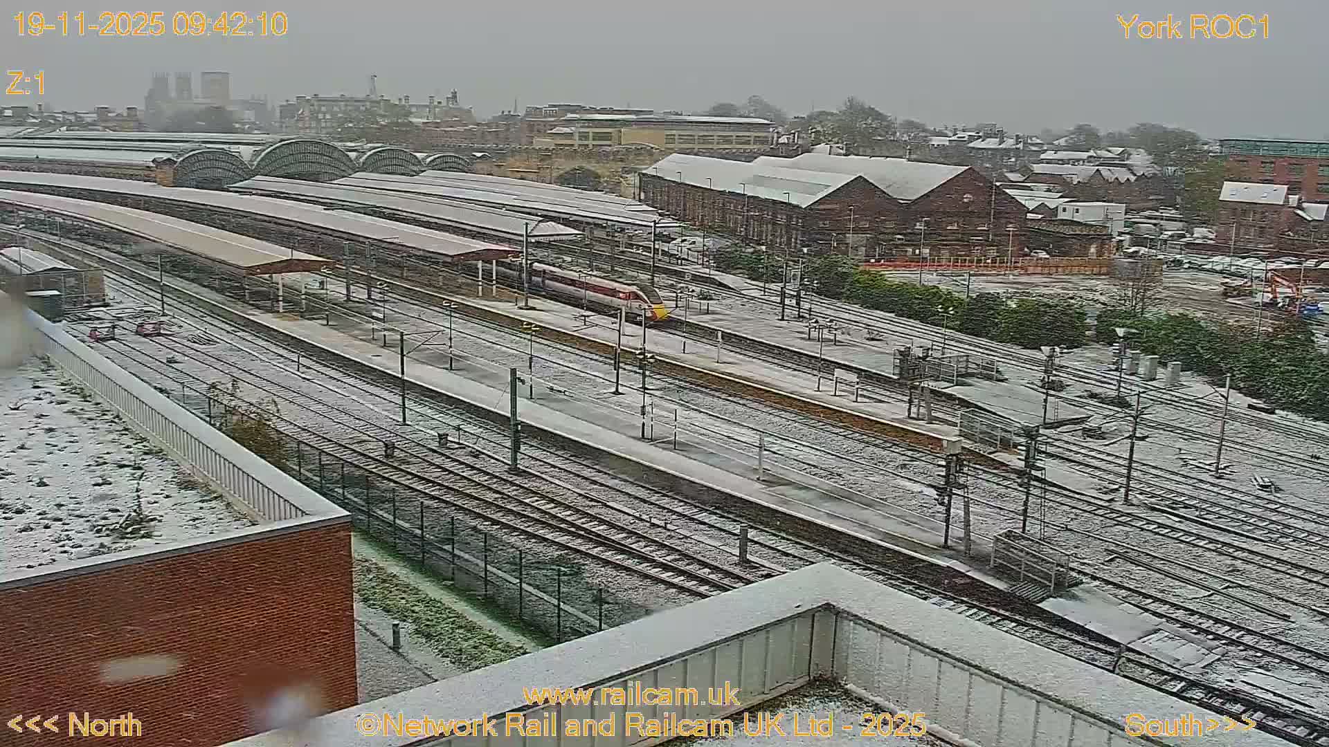 A broad view of a snow-covered multi-platform train station is seen on an overcast day, with a red and white train visible on one of the tracks.