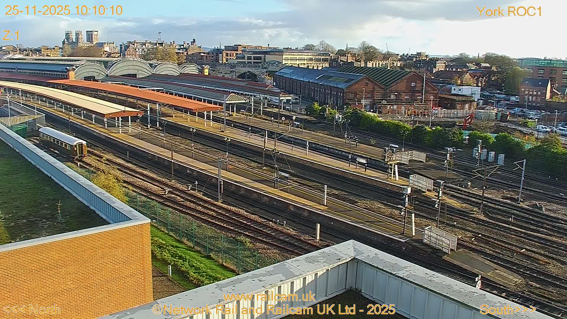 An elevated view captures a sprawling outdoor train station with numerous tracks and platforms, a lone train carriage, and an urban backdrop featuring diverse buildings and a prominent cathedral in the distance, all beneath a bright, partly cloudy sky.