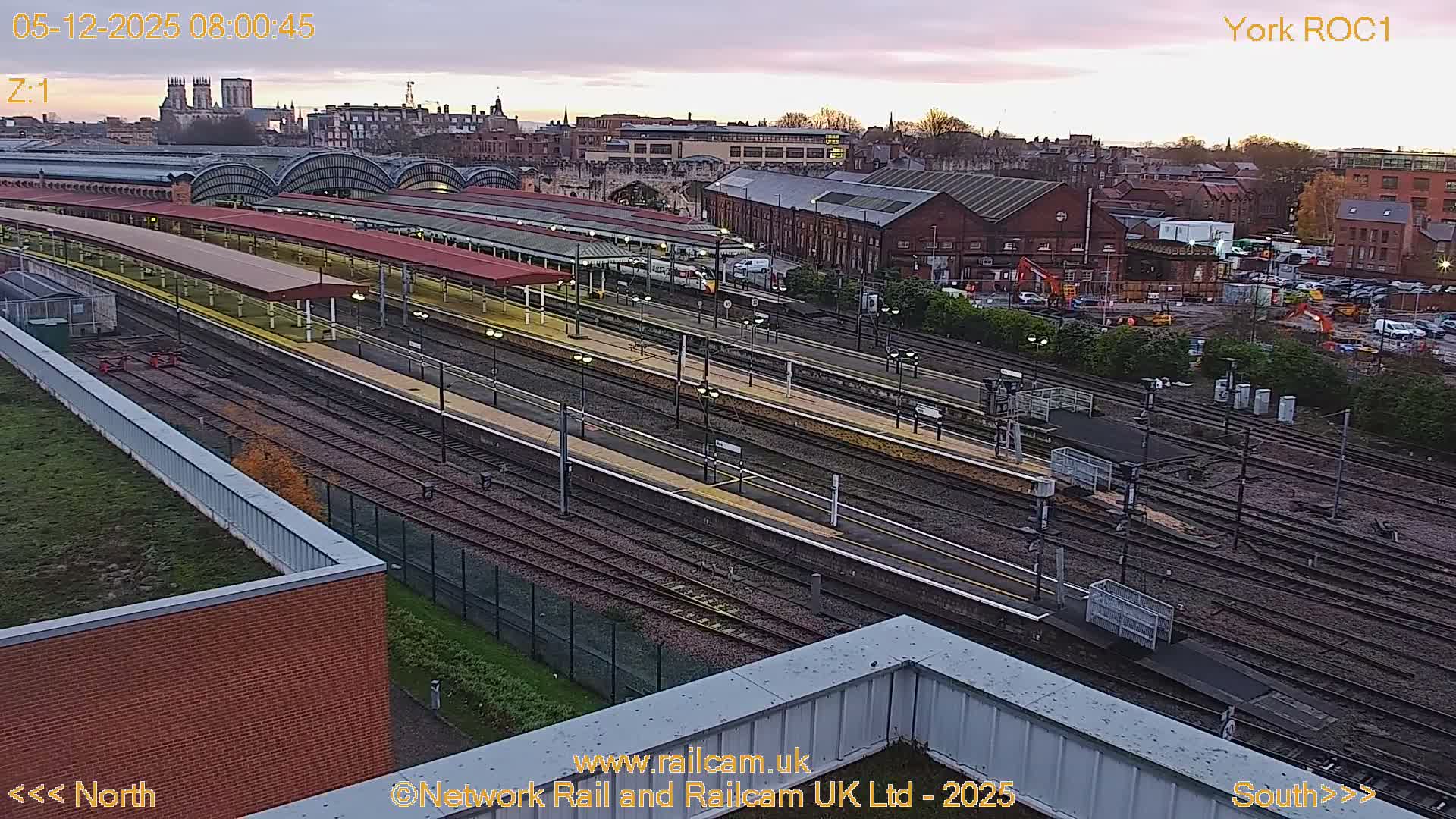 An elevated view captures a sprawling train station with multiple tracks and platforms, some illuminated under red canopies, set against a soft, cloudy dawn sky with distant city buildings including a prominent cathedral.
