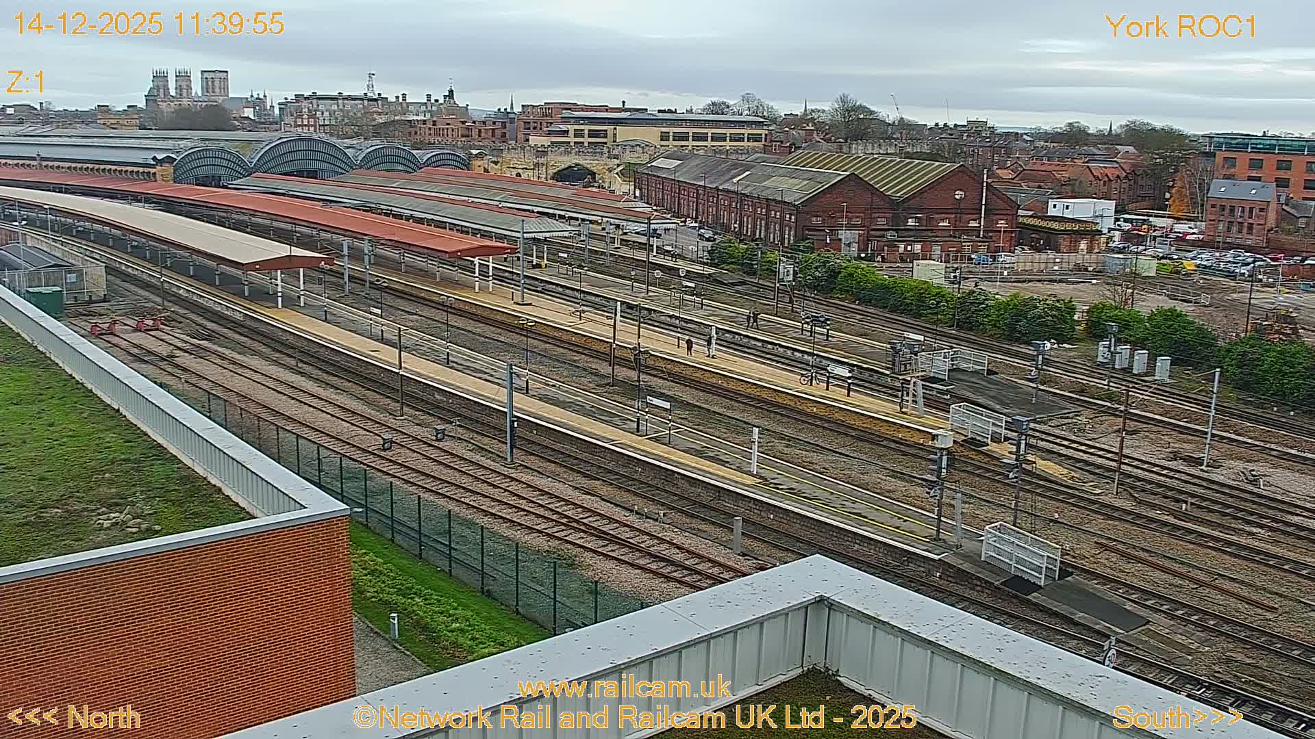 An elevated view captures a sprawling train station with multiple tracks and platforms, some illuminated under red canopies, set against a soft, cloudy dawn sky with distant city buildings including a prominent cathedral.