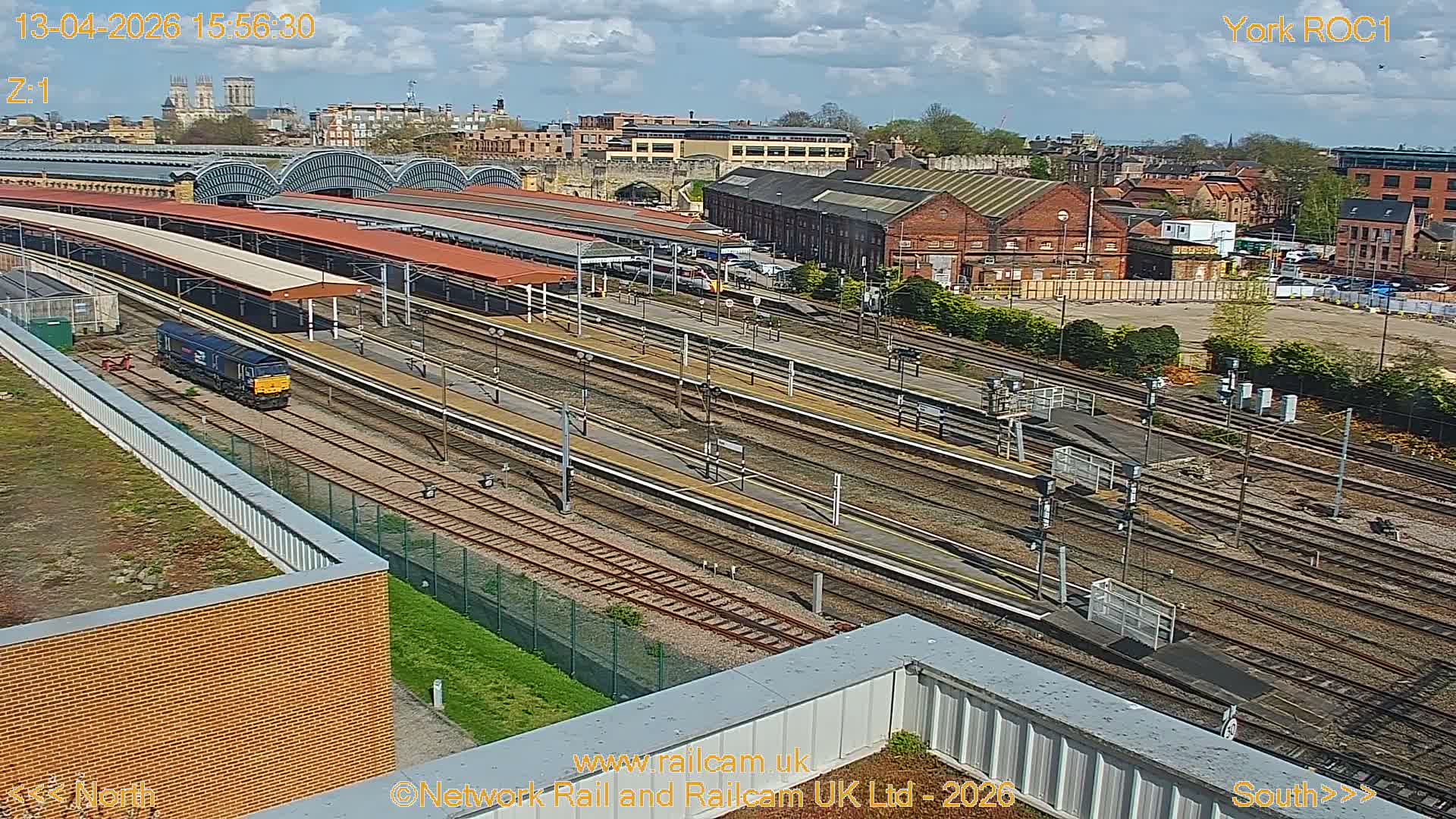An elevated view captures a sprawling train station with multiple tracks and platforms, some illuminated under red canopies, set against a soft, cloudy dawn sky with distant city buildings including a prominent cathedral.