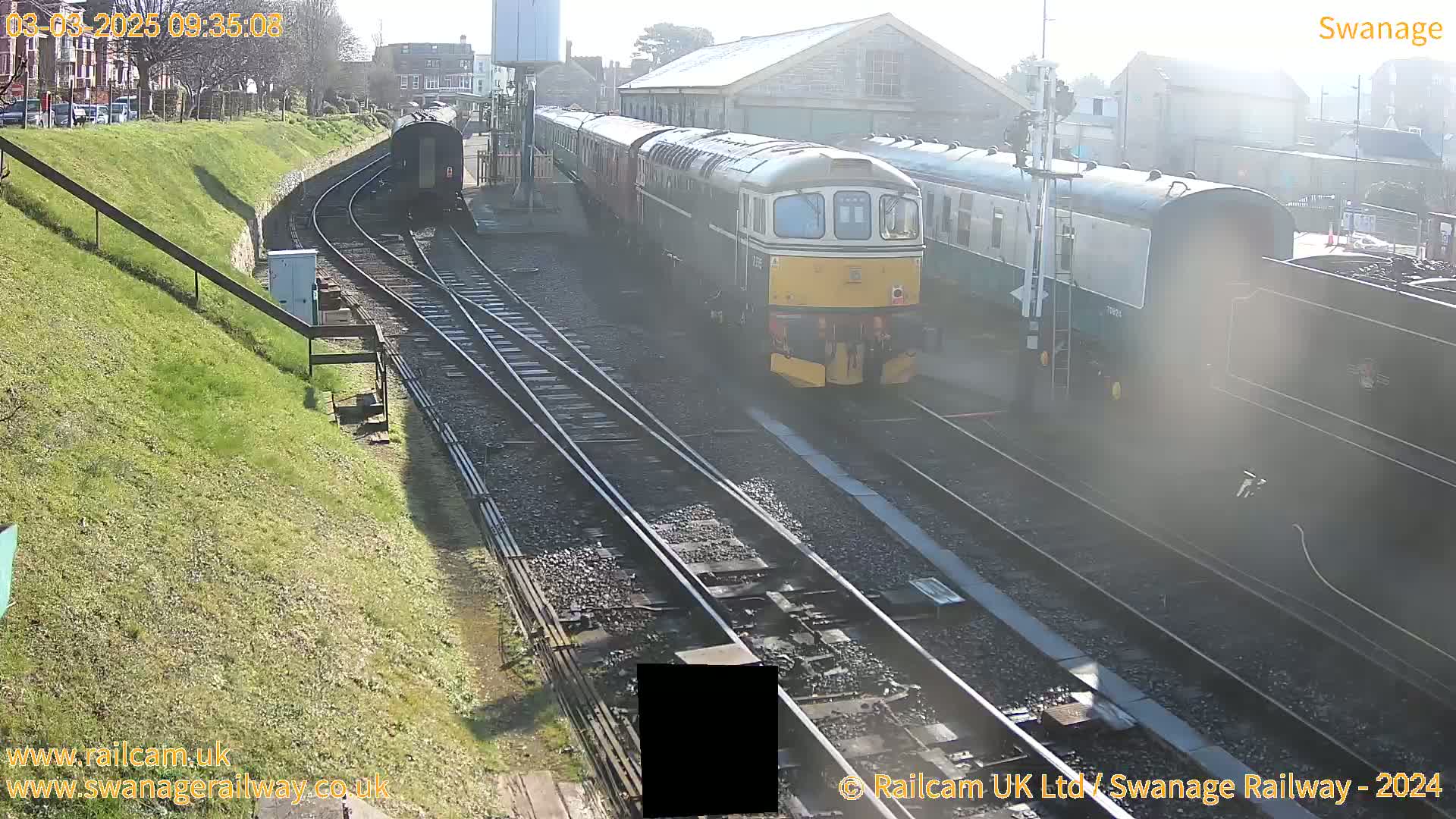 A diesel locomotive leads a train of passenger cars on a railway track on a sunny day.