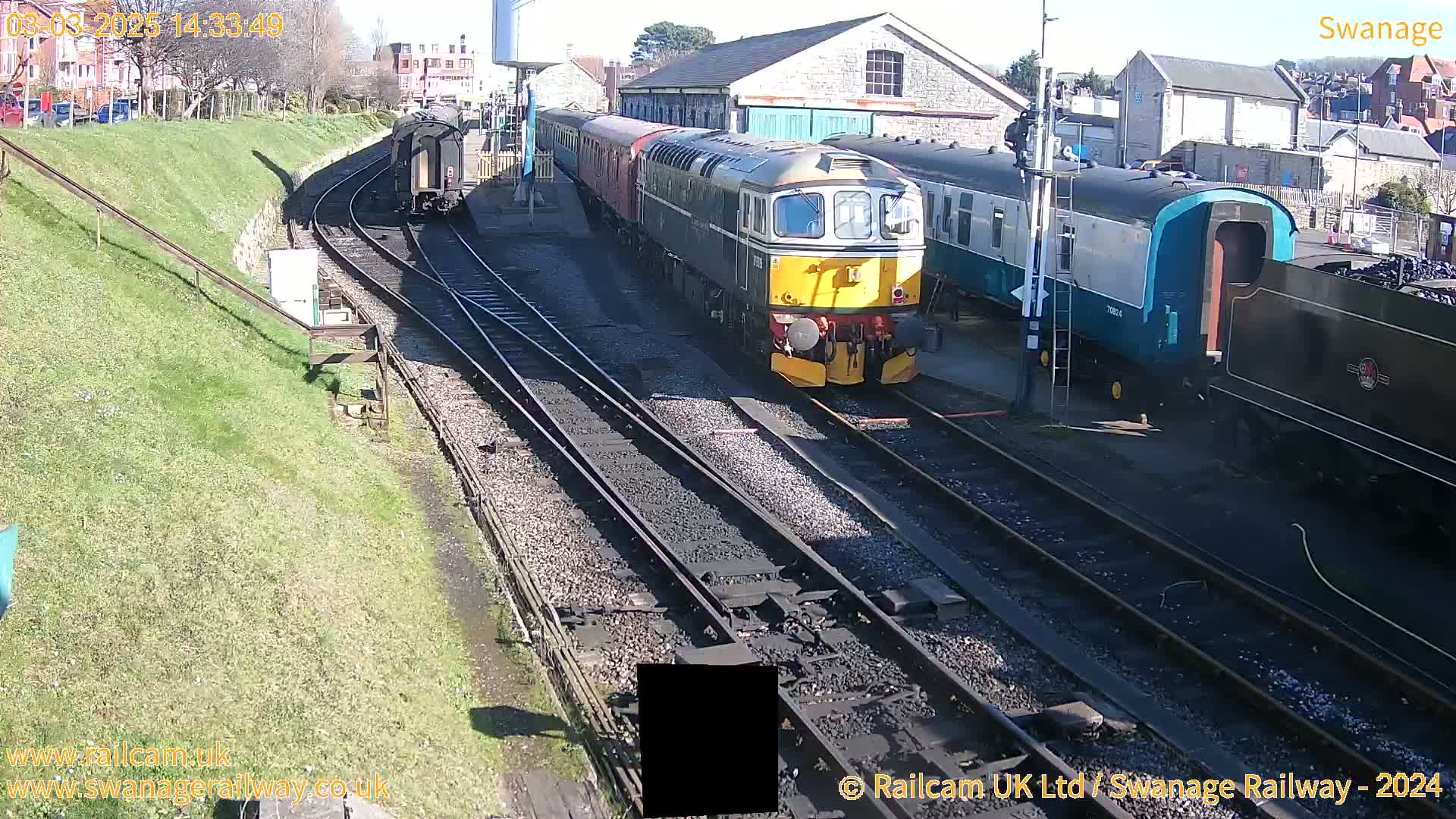 A sunny day at a railway station shows several train cars and a diesel locomotive on the tracks, with a grassy embankment and buildings in the background.