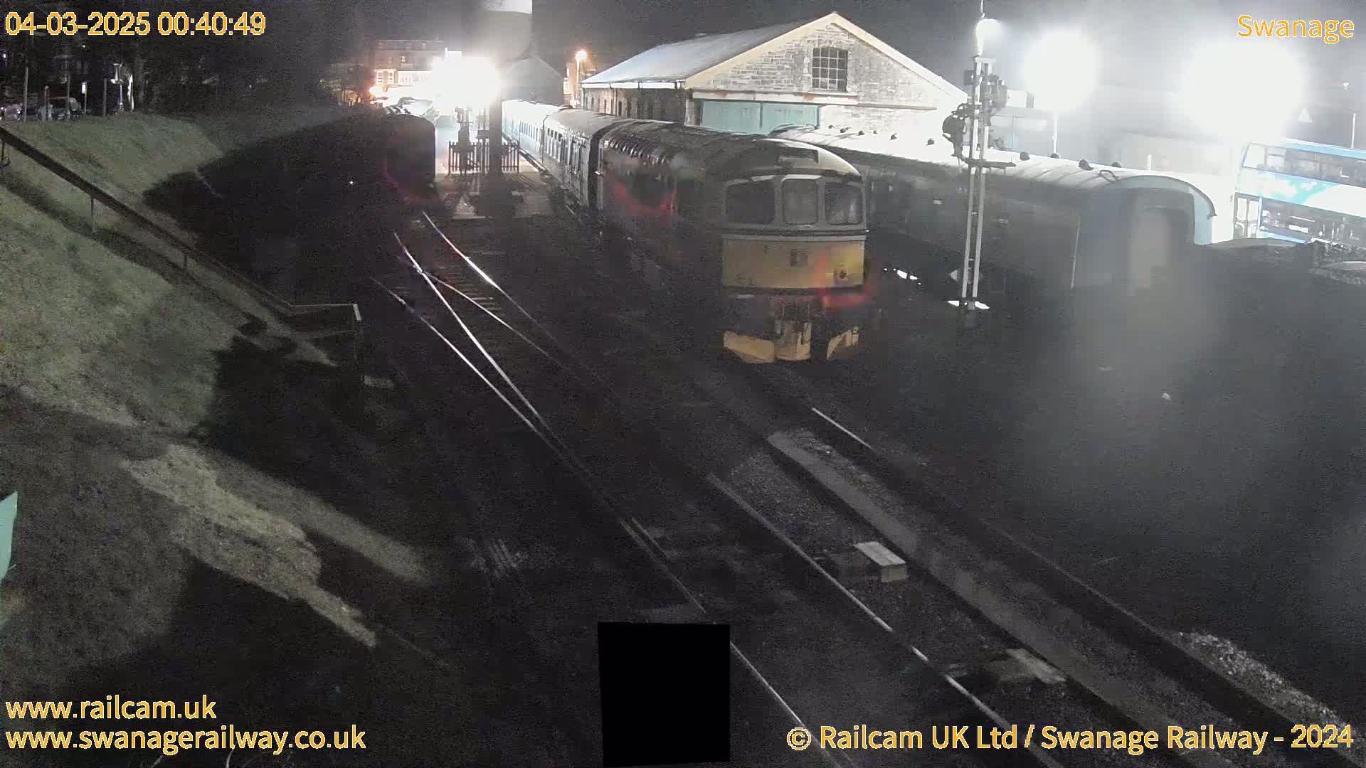 A train sits at a nighttime railway station under artificial light, next to another train and a bus.