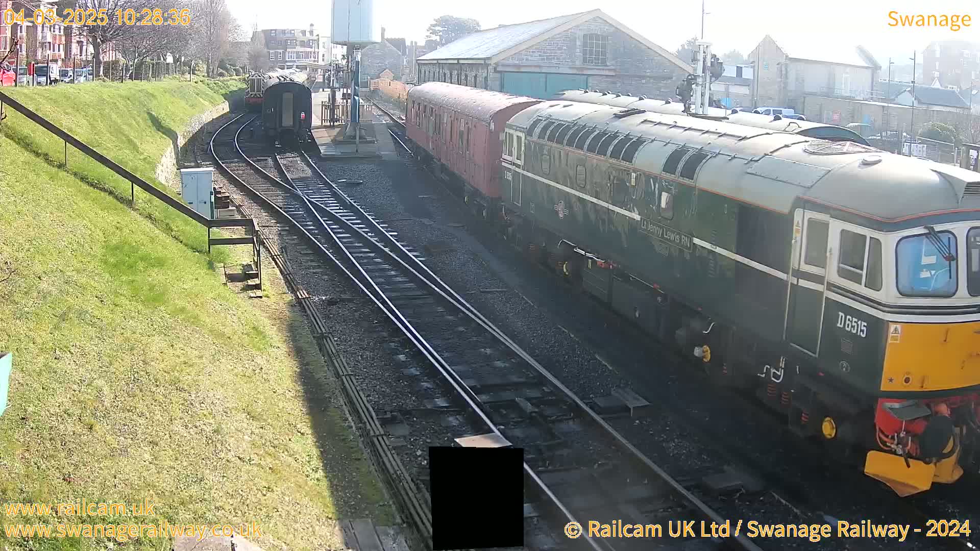A green diesel locomotive pulls a line of red passenger cars along railway tracks on a sunny day.