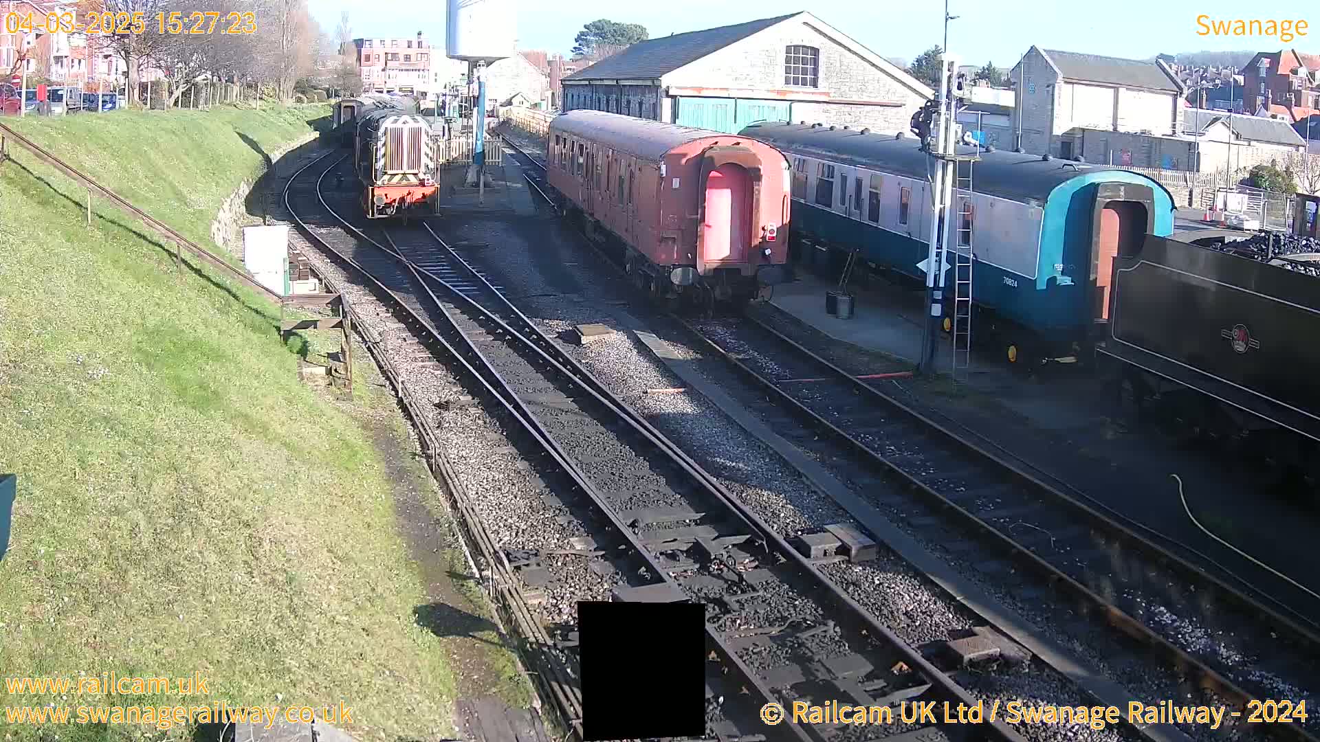 Several train cars and locomotives are parked on railway tracks on a sunny day near a stone building.