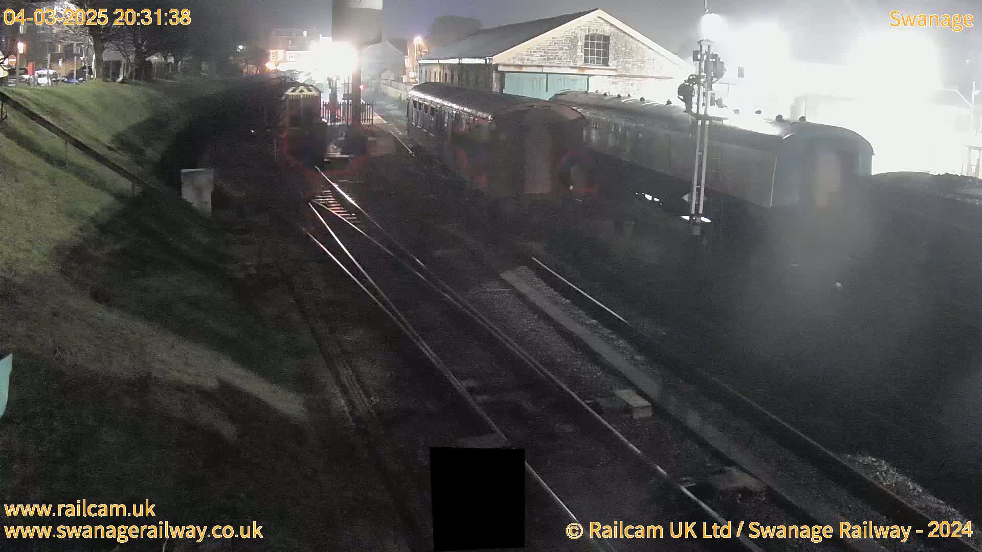 A train sits at night in a railway station next to a stone building under overcast skies.