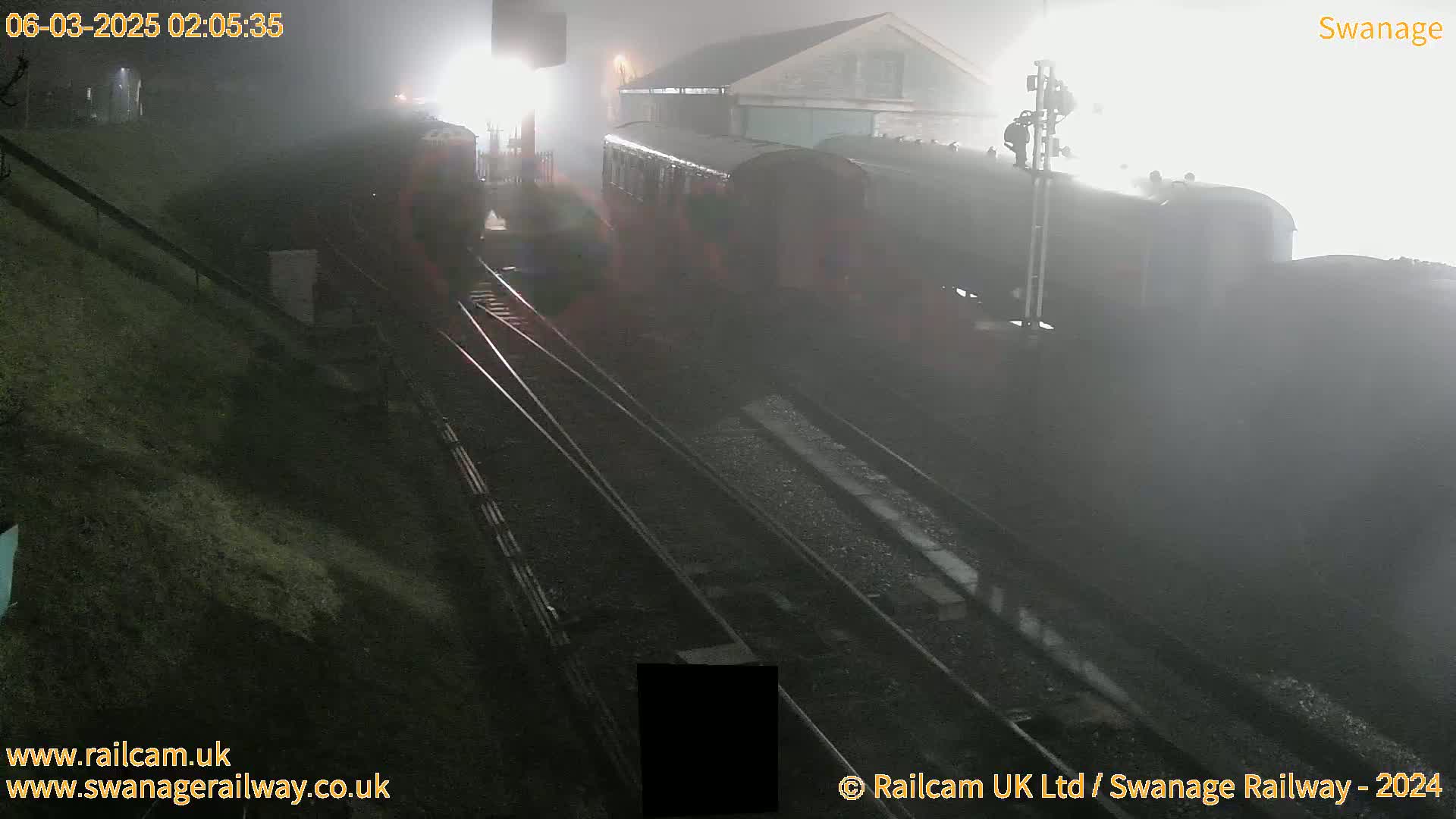 A dark-colored train sits in a foggy nighttime railway station next to a building.
