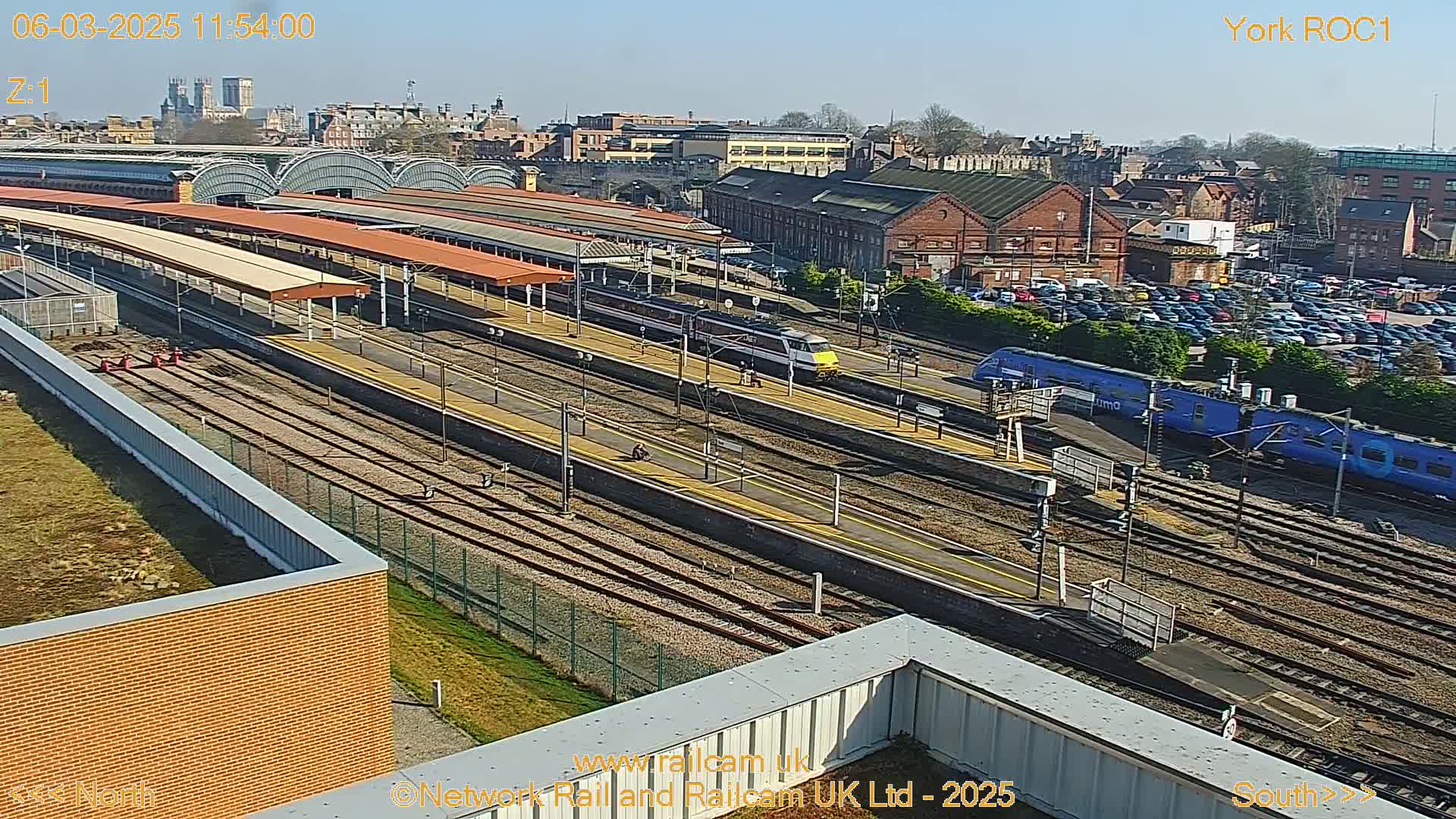 A high-angle view of a train station on a sunny day shows multiple train tracks with several trains present, along with a large parking lot and a cityscape in the background.