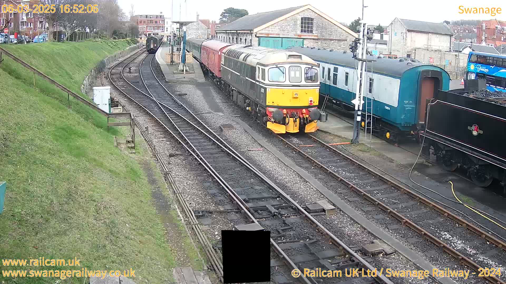 A diesel locomotive and several railway carriages are parked on train tracks next to a railway station building on a partly cloudy day.