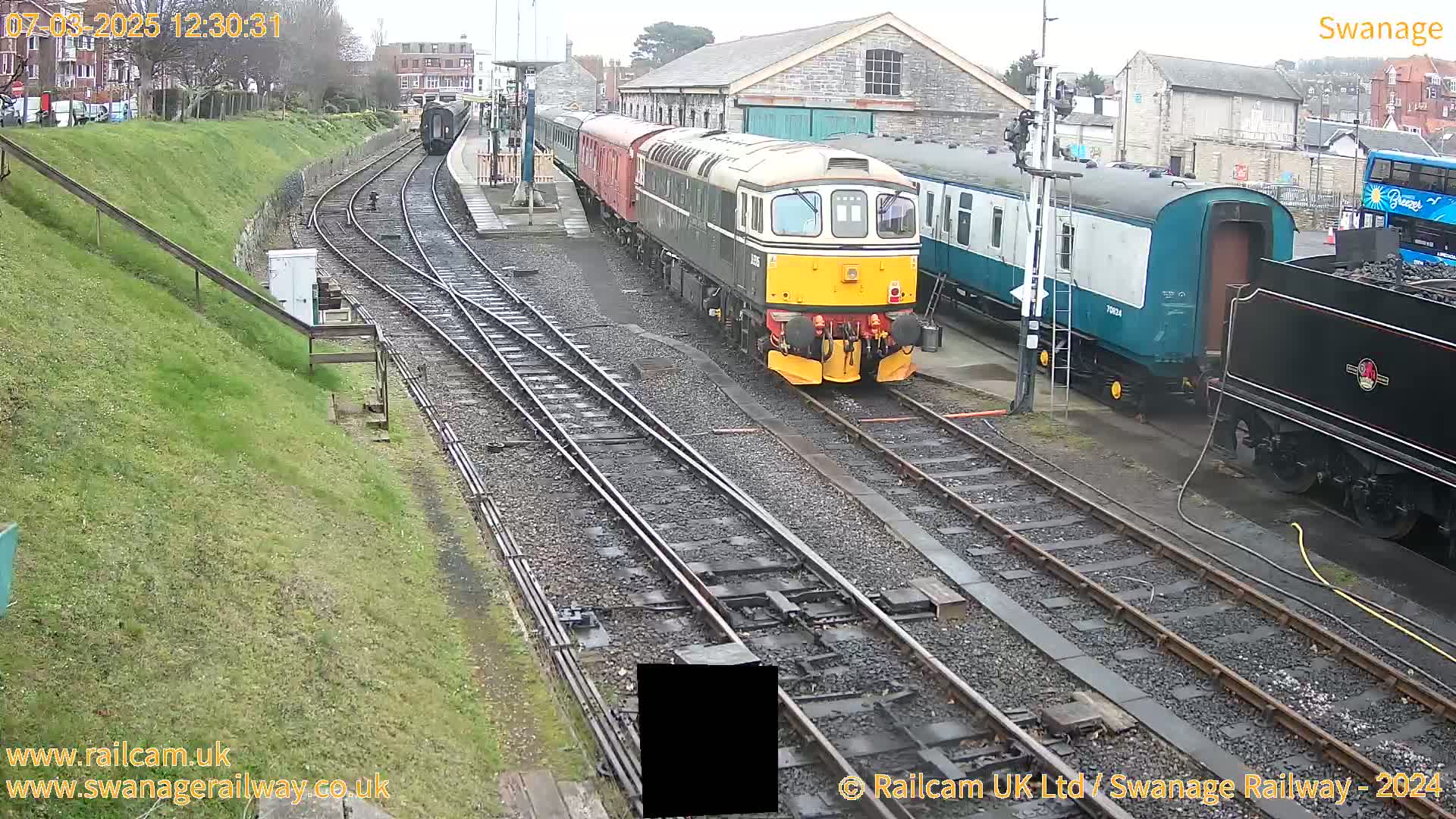 A diesel locomotive leads a passenger train past other railcars and a steam locomotive at a railway station on an overcast day.