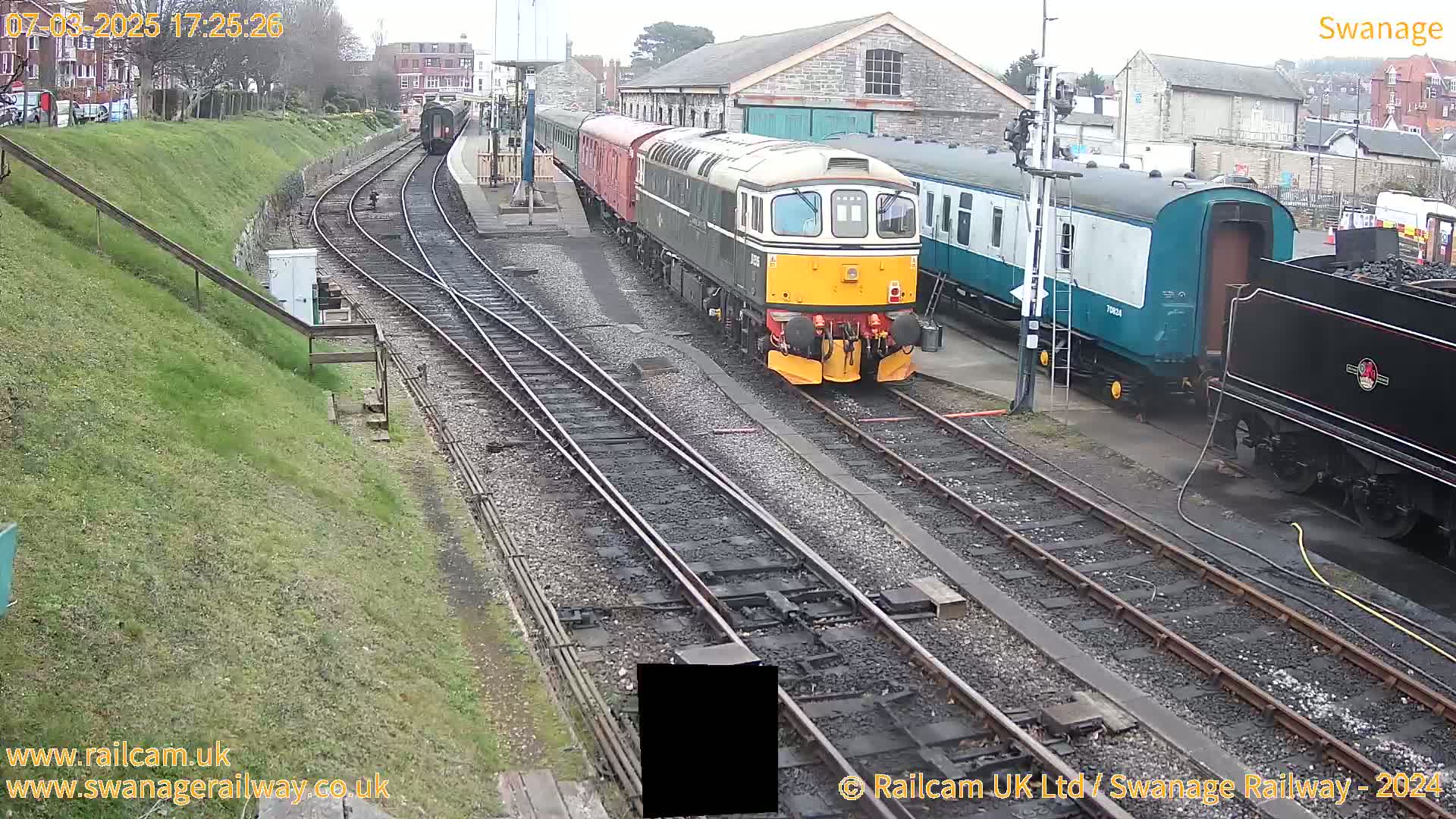 A diesel locomotive pulls a train of passenger cars past a station on an overcast day, with other trains and railway buildings nearby.