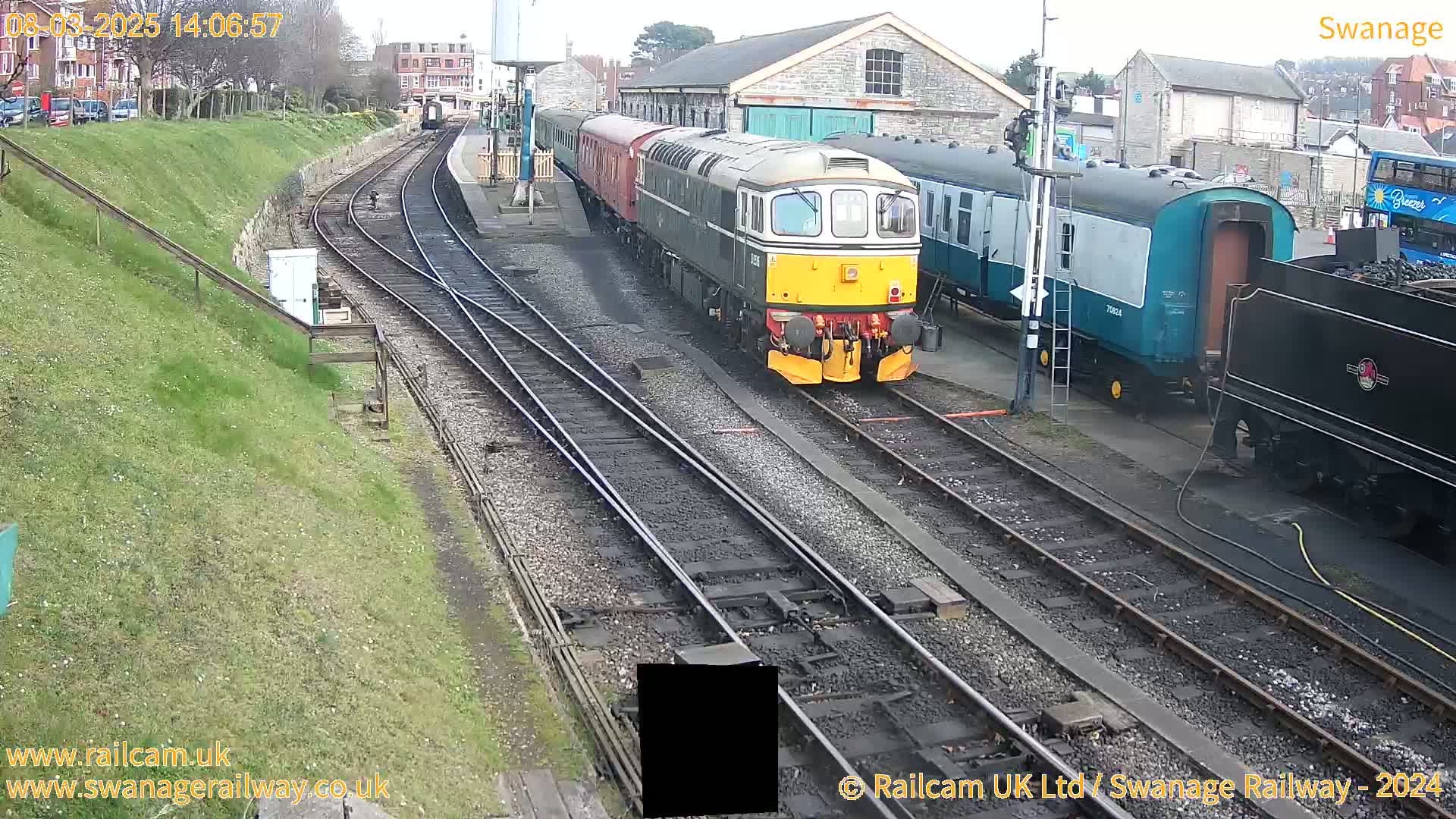 A diesel locomotive leads a passenger train past a railway station on a partly sunny day.