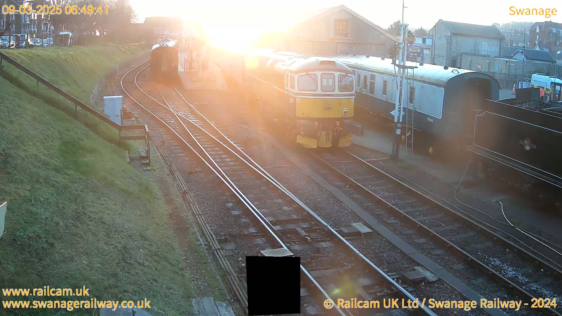 A train sits on railroad tracks at sunrise, with other railway cars and buildings visible in the background.