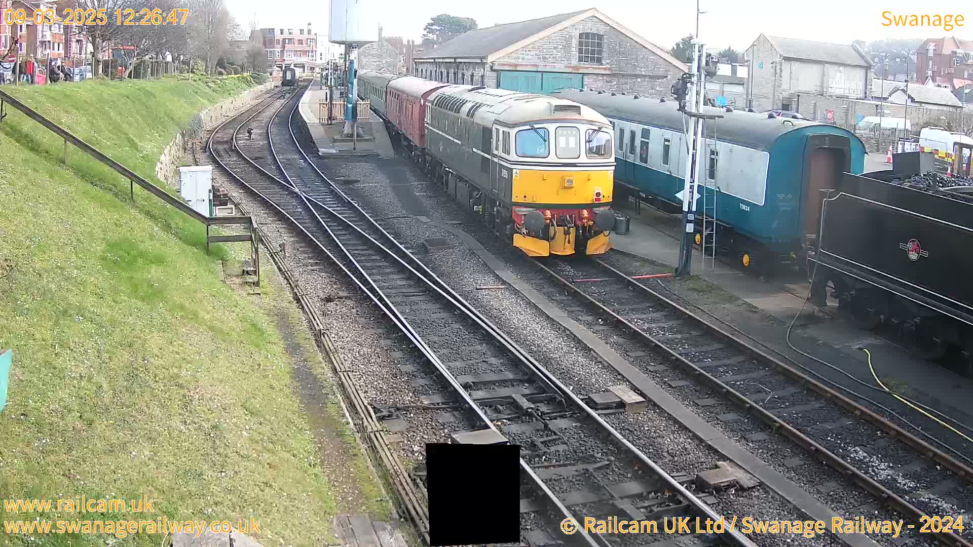 A diesel locomotive and several railway carriages are parked on train tracks next to a station building on a partly sunny day.