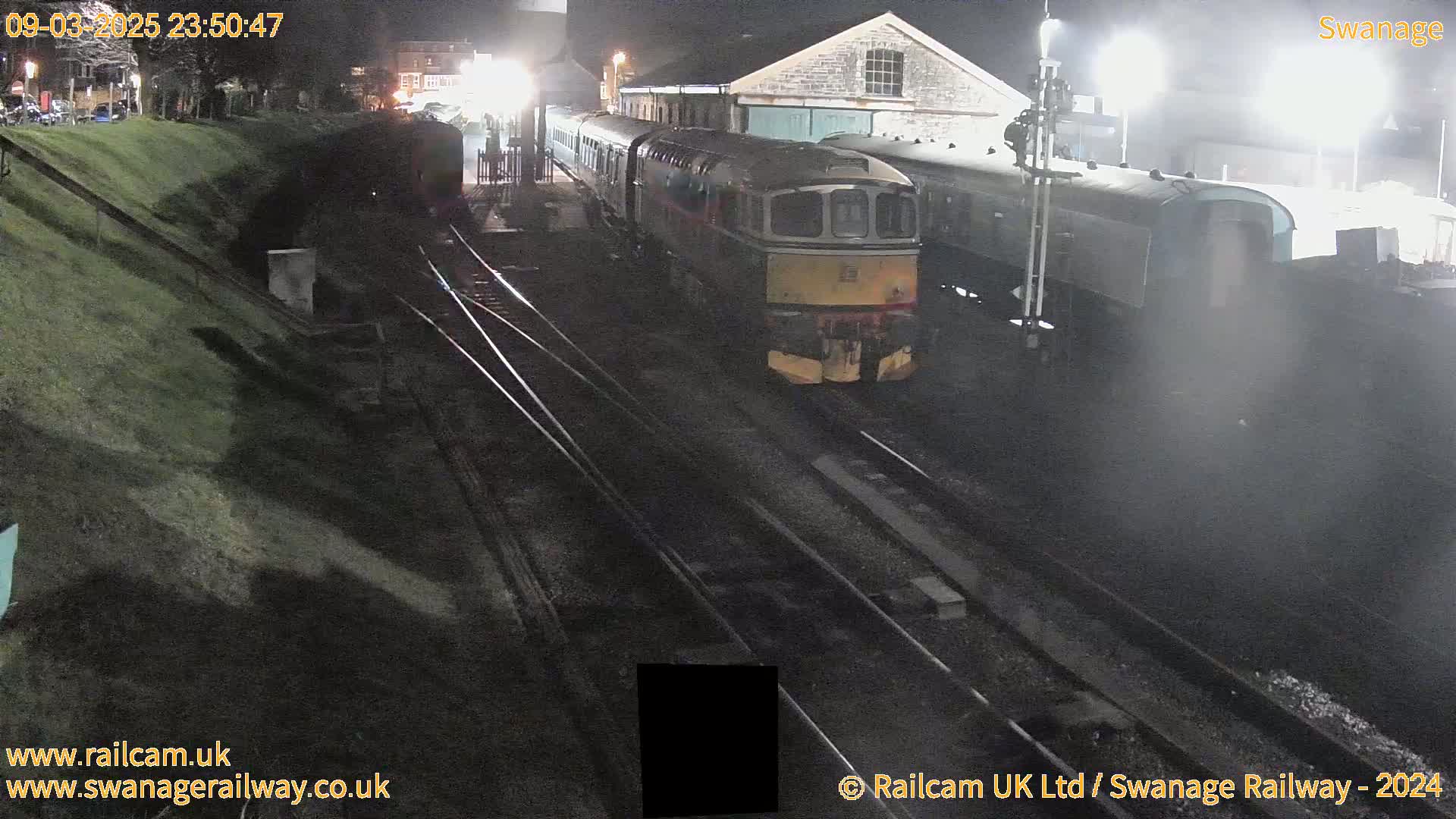 A diesel locomotive and passenger cars are parked at a nighttime train station next to a stone building.