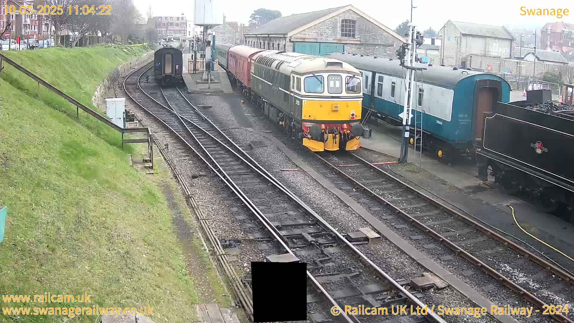 A diesel locomotive and several railway carriages are parked on train tracks beside a grassy embankment on an overcast day.