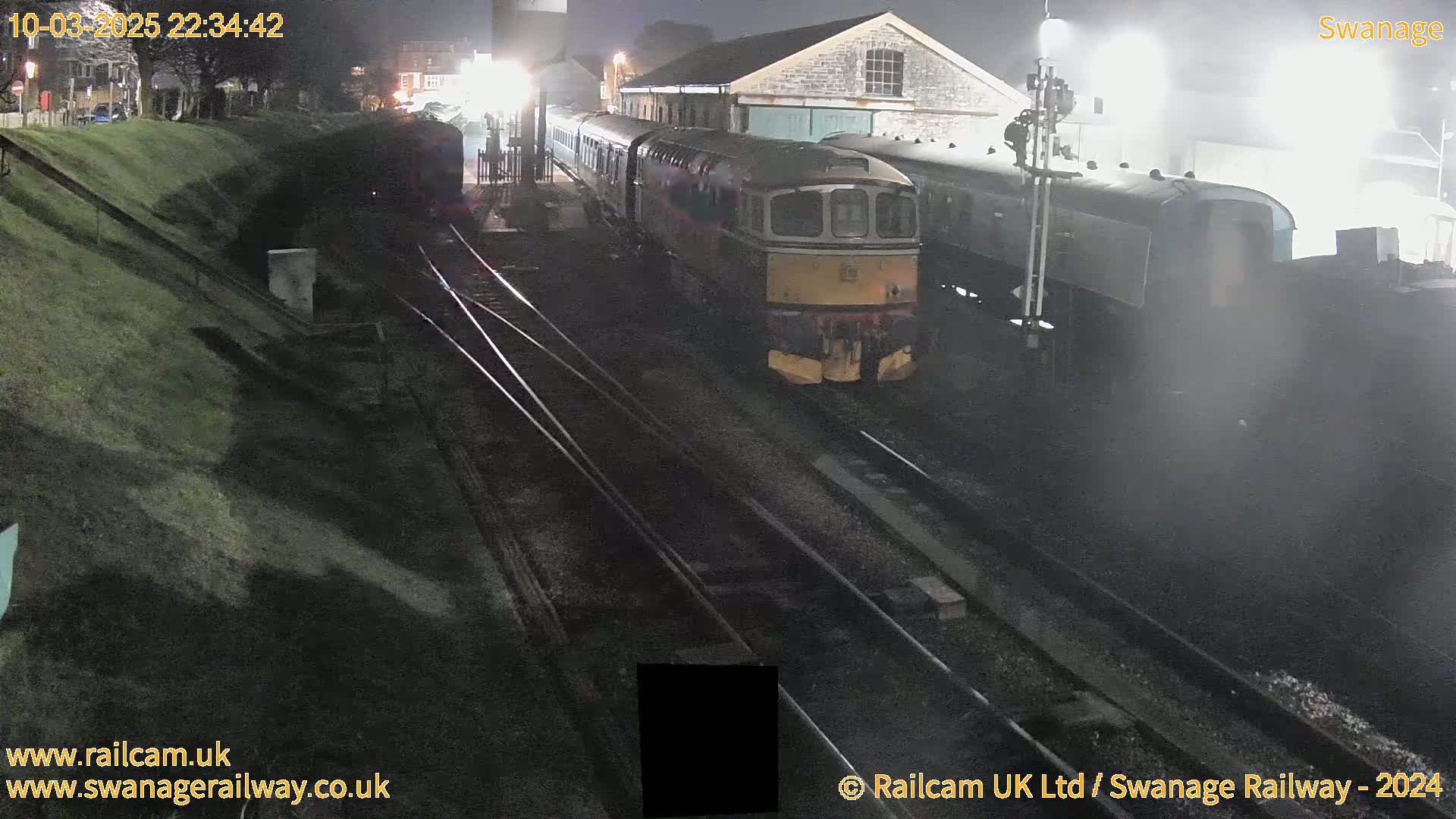 A diesel train is parked at night next to a stone building at a train station.