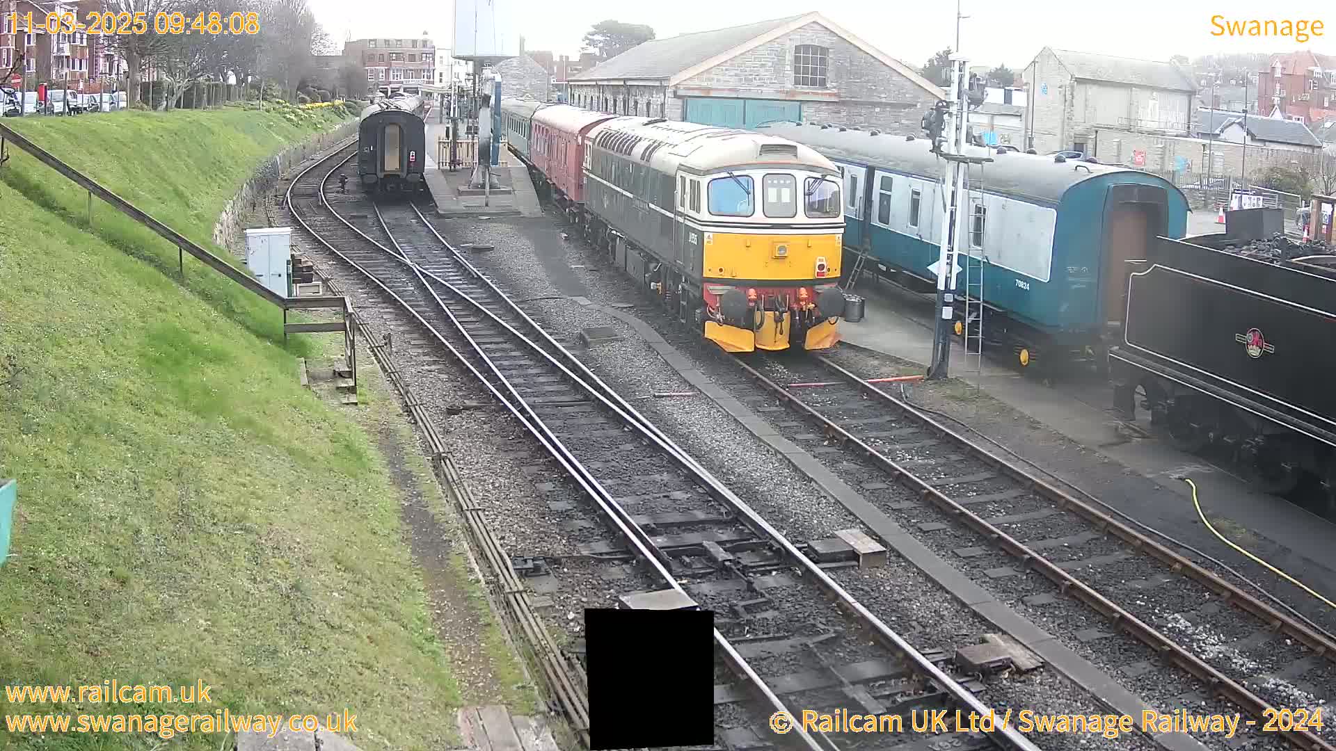 A diesel locomotive and several passenger cars are parked on railway tracks next to a train station on an overcast day.