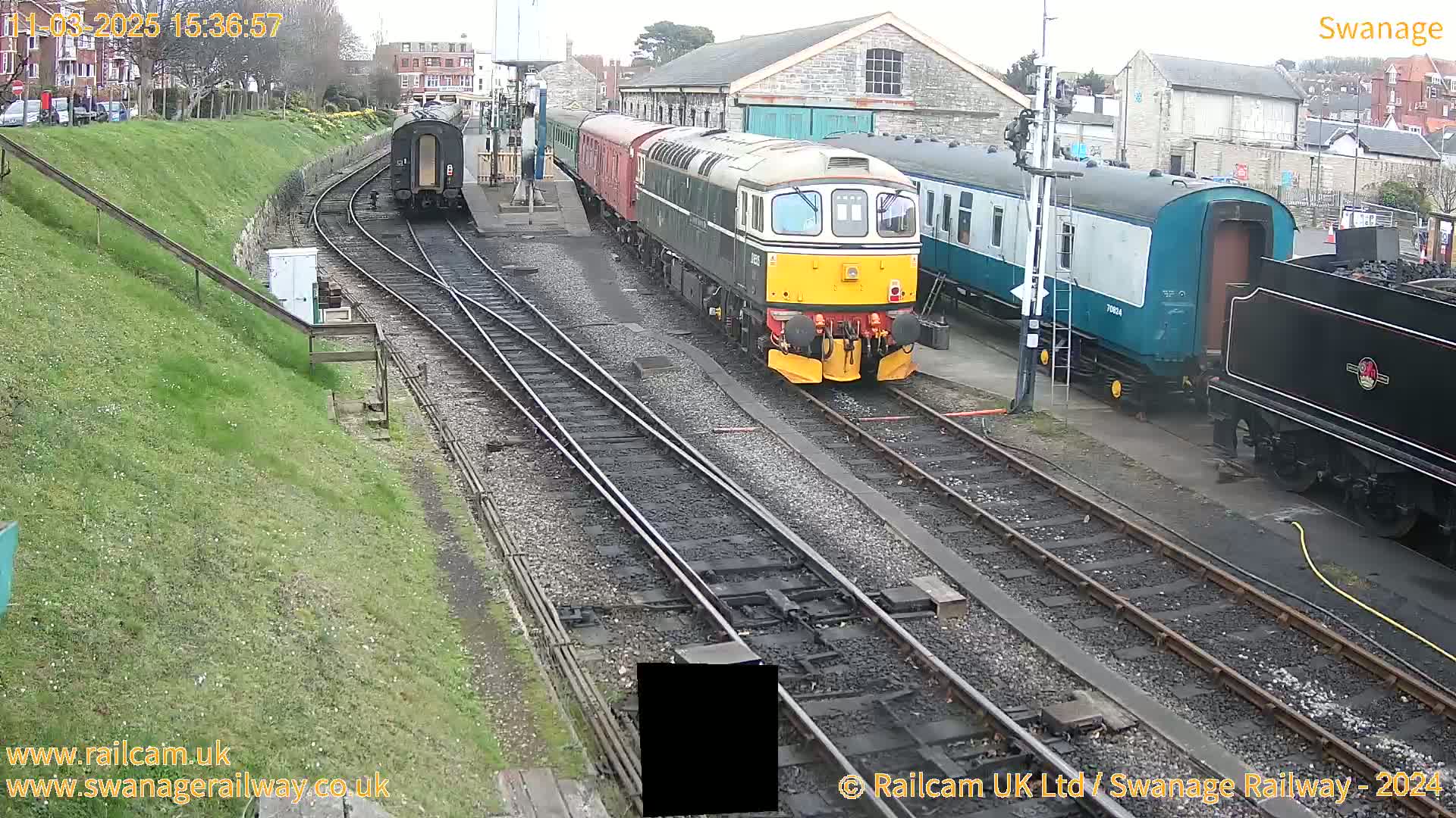 Several trains, including a diesel locomotive and passenger cars, are parked on railway tracks in a train yard on an overcast day.