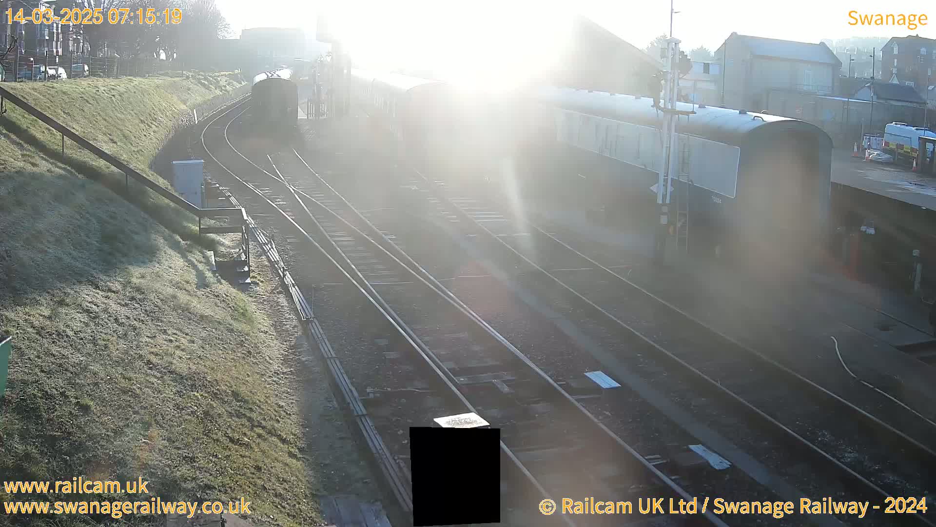 A hazy sunrise illuminates a railway station with several train cars parked on the tracks, alongside a grassy embankment.