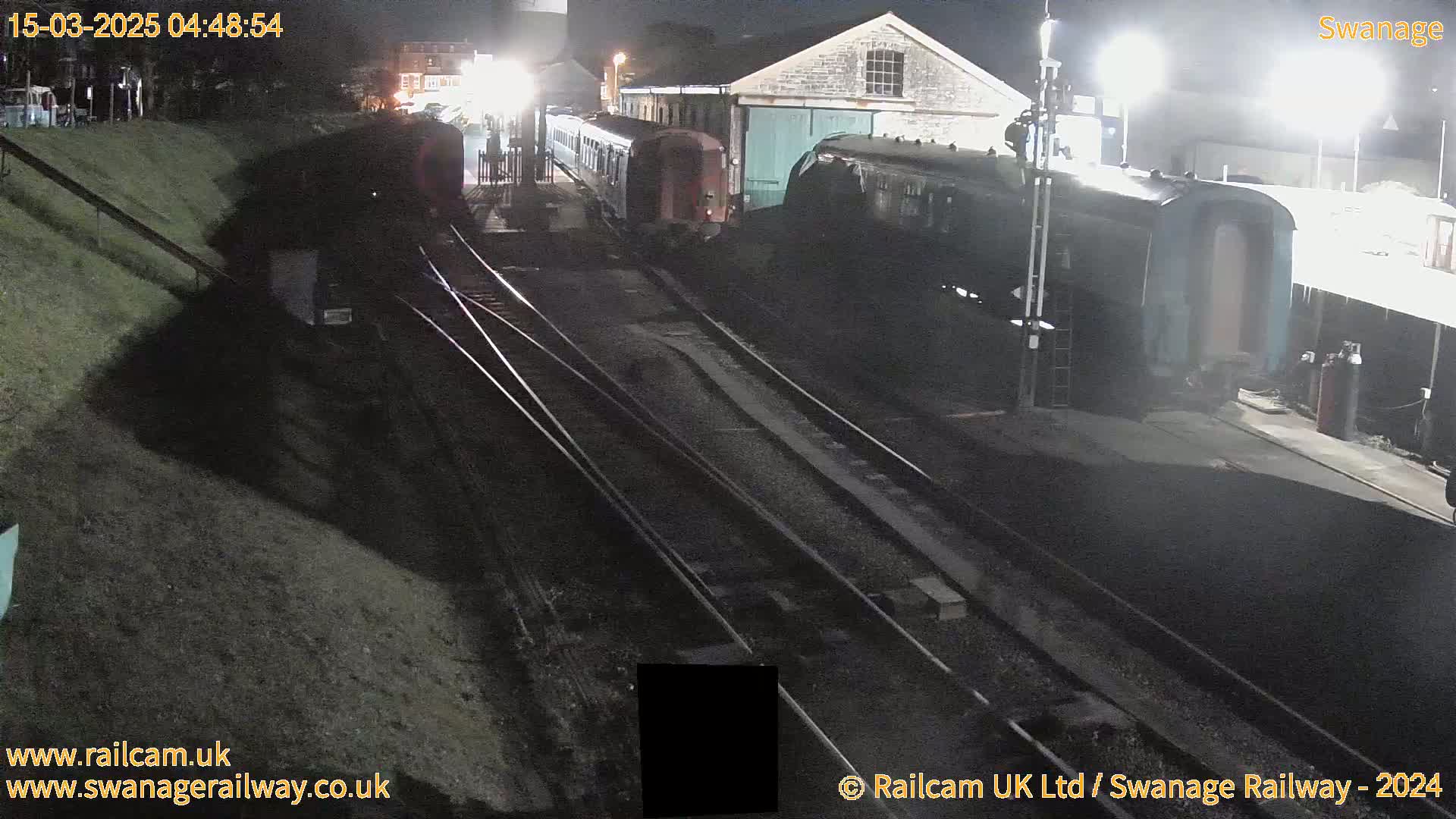 At night, several train cars are parked on railway tracks next to a building, under artificial lighting.