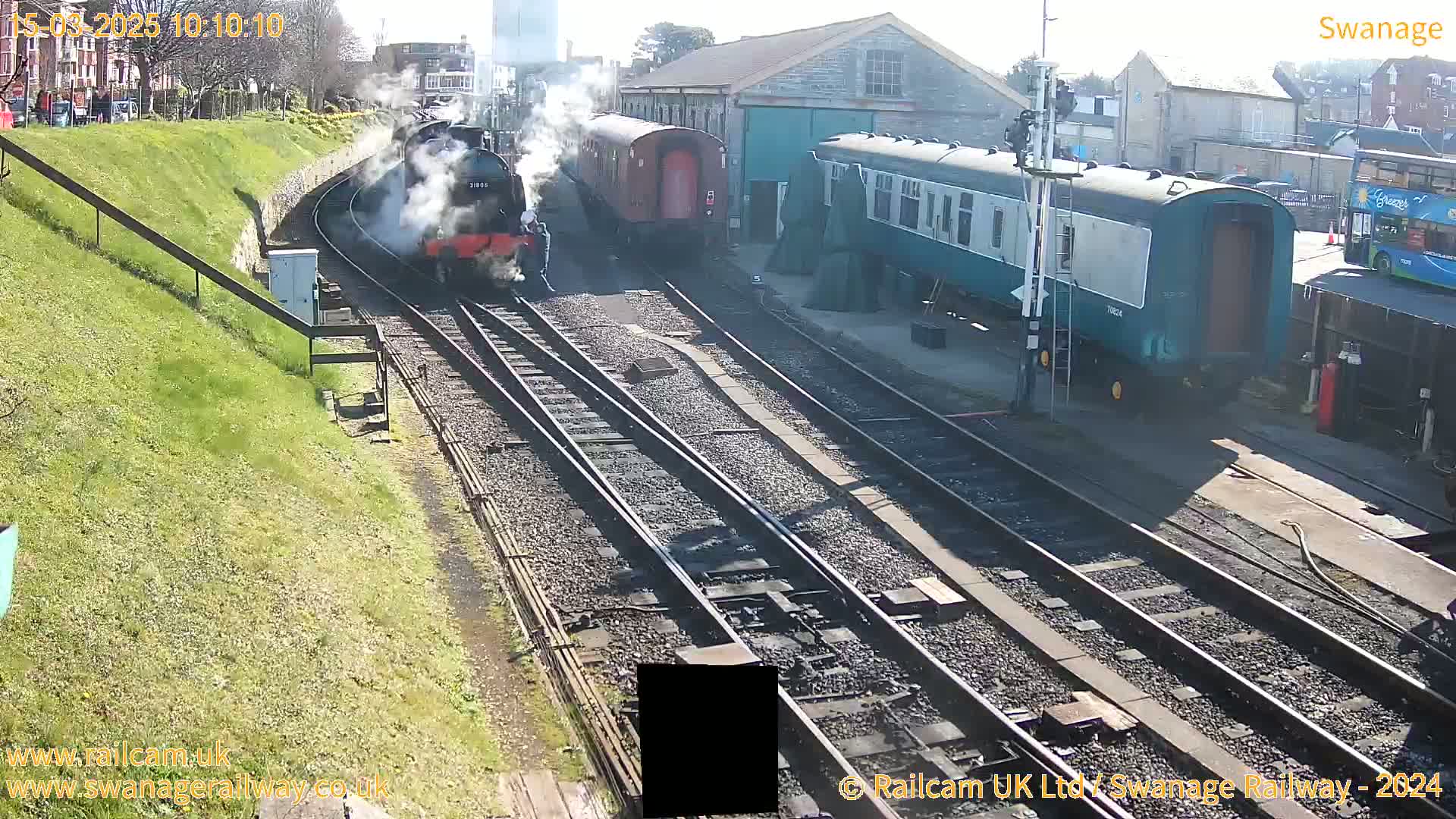 A steam train pulls a line of carriages past a railway station on a sunny day.