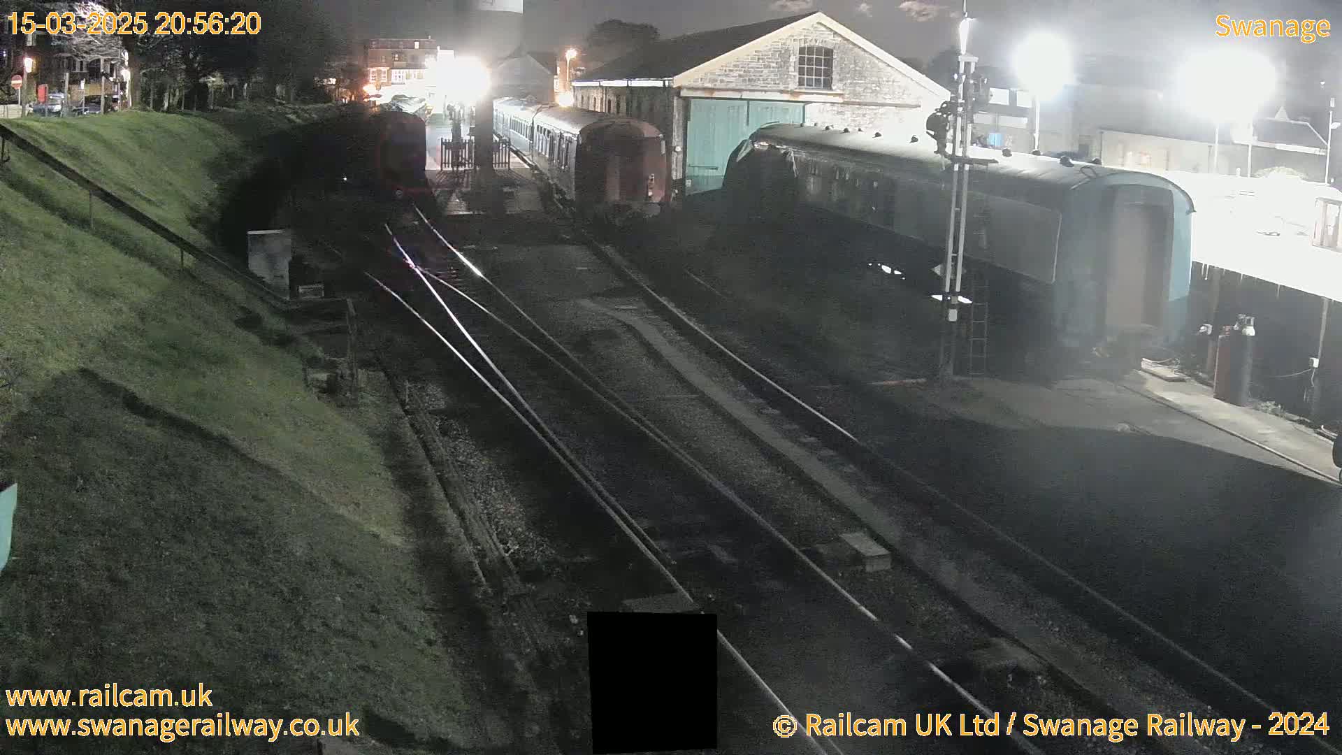 Several railway carriages are parked at night on railway tracks next to a stone building, under bright artificial lights.