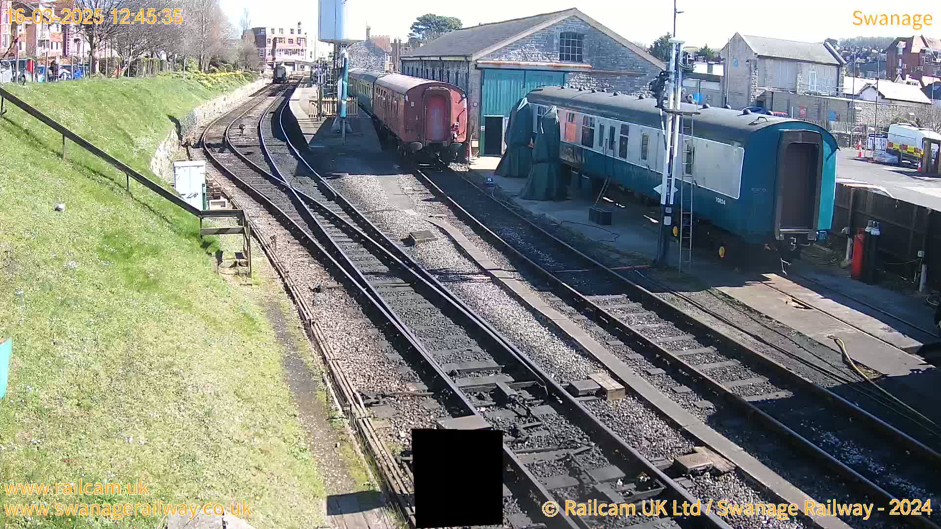 Railway tracks and several railway carriages, some parked and others appearing to be in transit, are visible on a sunny day.