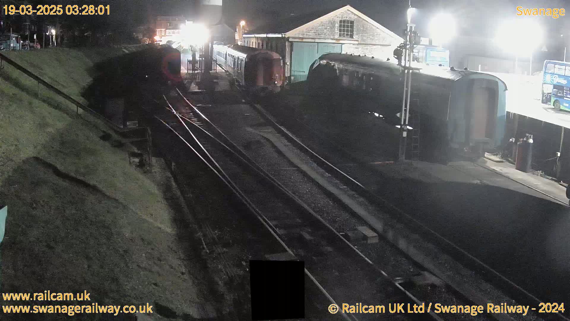 At night, a train is parked at a railway station near a building and other railway cars, under bright lights.