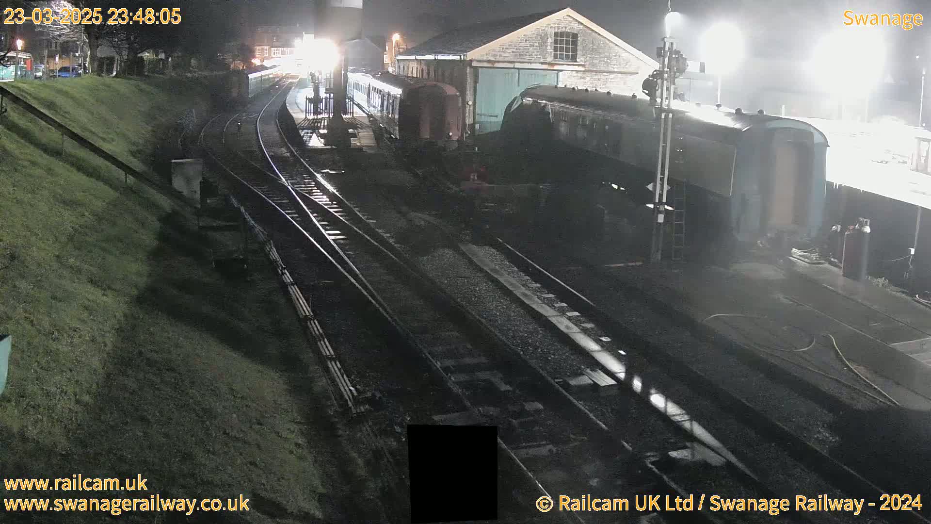 At night, a train sits in a rail yard next to a stone building, under bright lights and some mist.