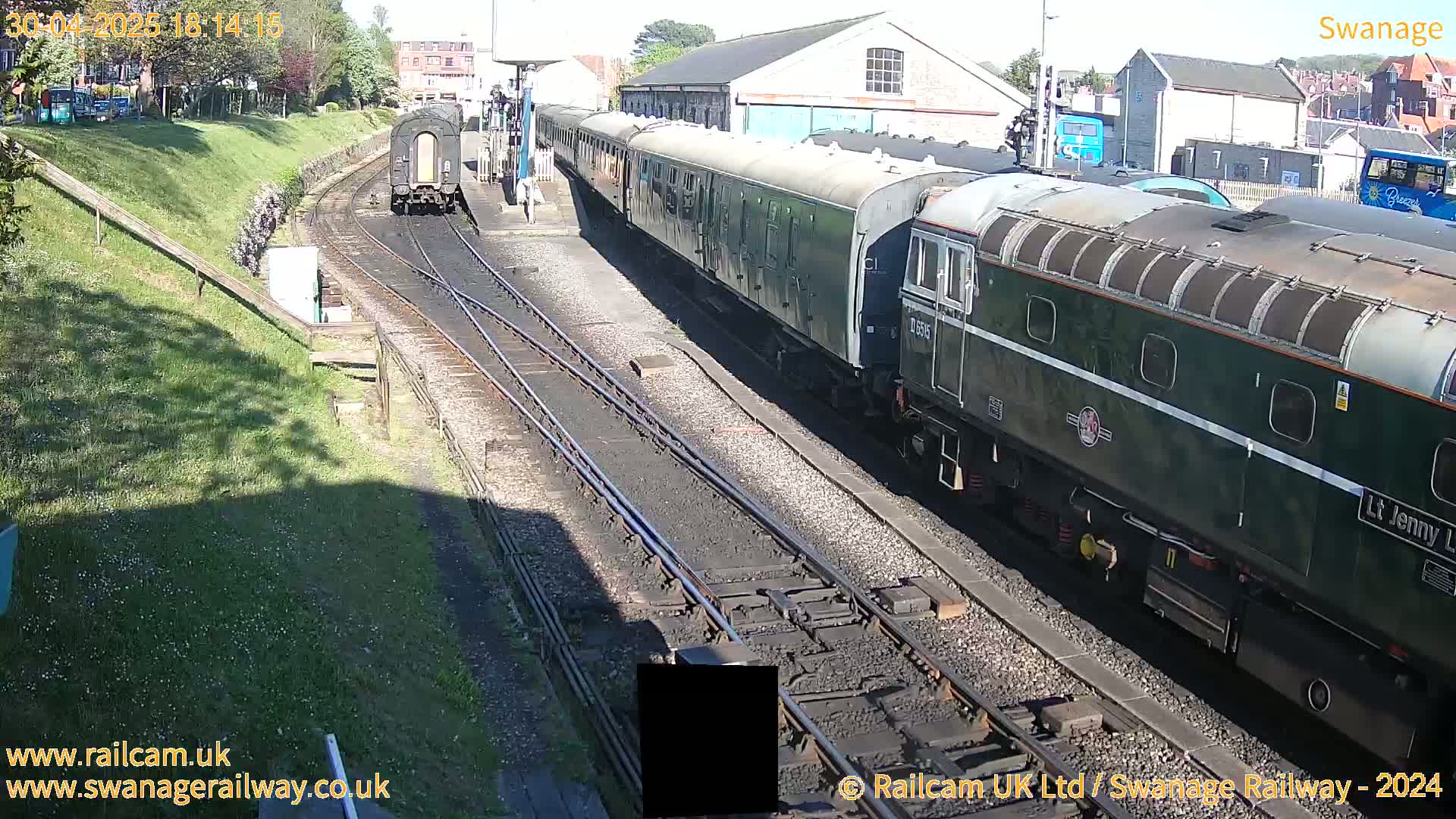 A long, dark-green passenger train is arriving at a railway station on a sunny day.
