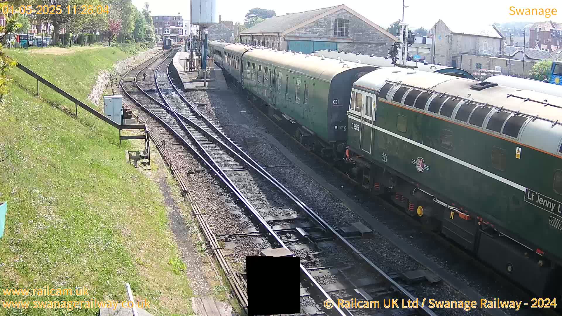 A dark green diesel locomotive and several passenger cars are parked on railway tracks on a sunny day.