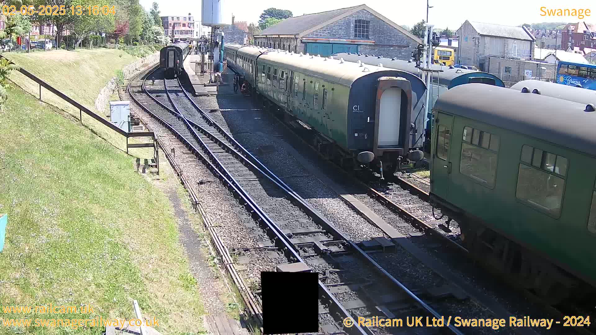 Several green railway carriages are parked on train tracks beside a grassy hill on a sunny day.