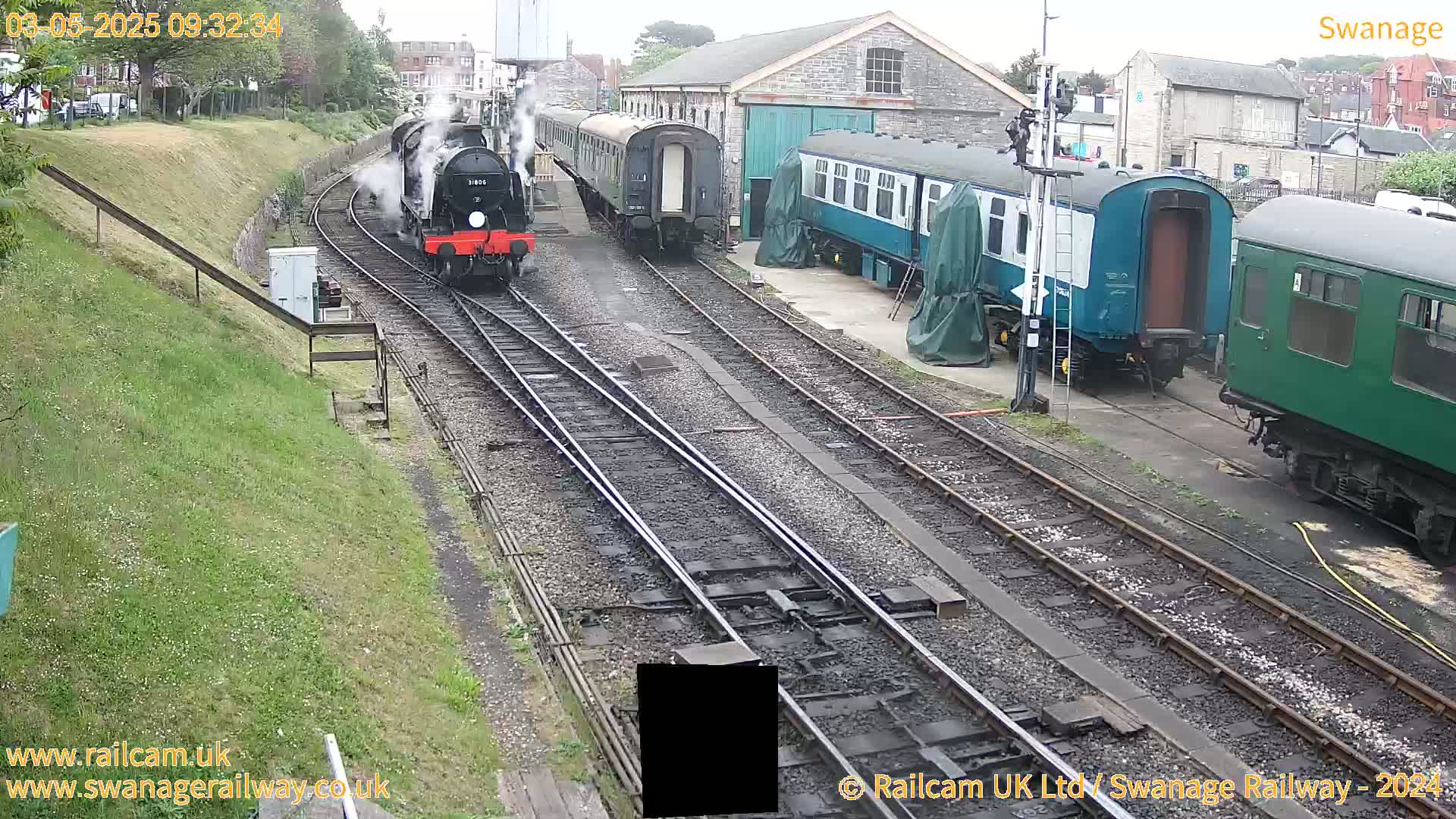 A steam train is moving along train tracks past railway cars and a depot on an overcast day.