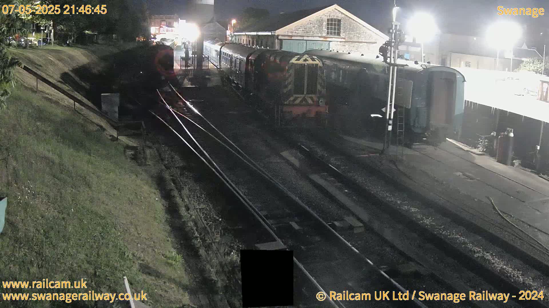 A diesel train sits at night next to a railway station and tracks.