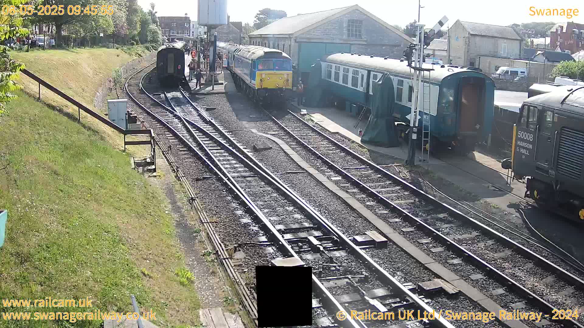 Several train cars and locomotives are parked on sunny railway tracks next to a stone building.