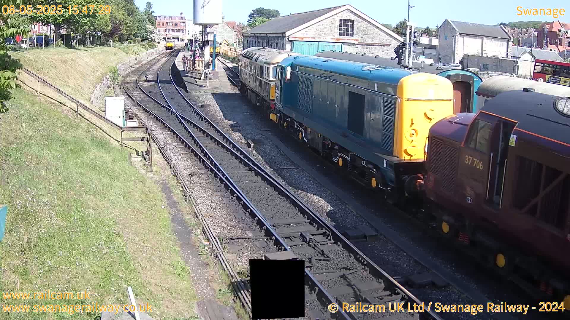 Multiple diesel locomotives are parked on railway tracks in bright sunlight next to a railway station building.