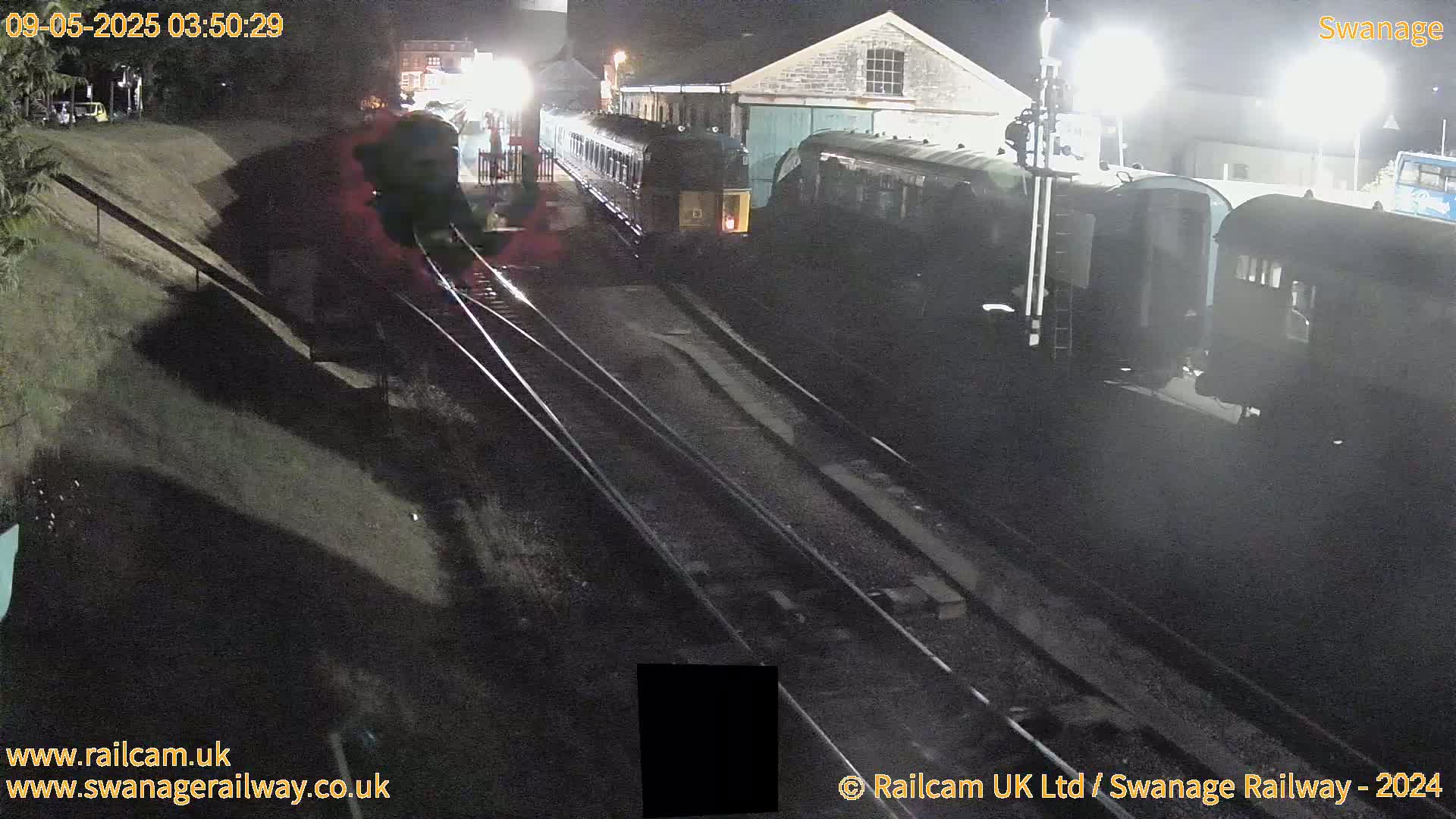 Several dark-colored railway cars are parked at night near a building, on railway tracks beside a sloped embankment.