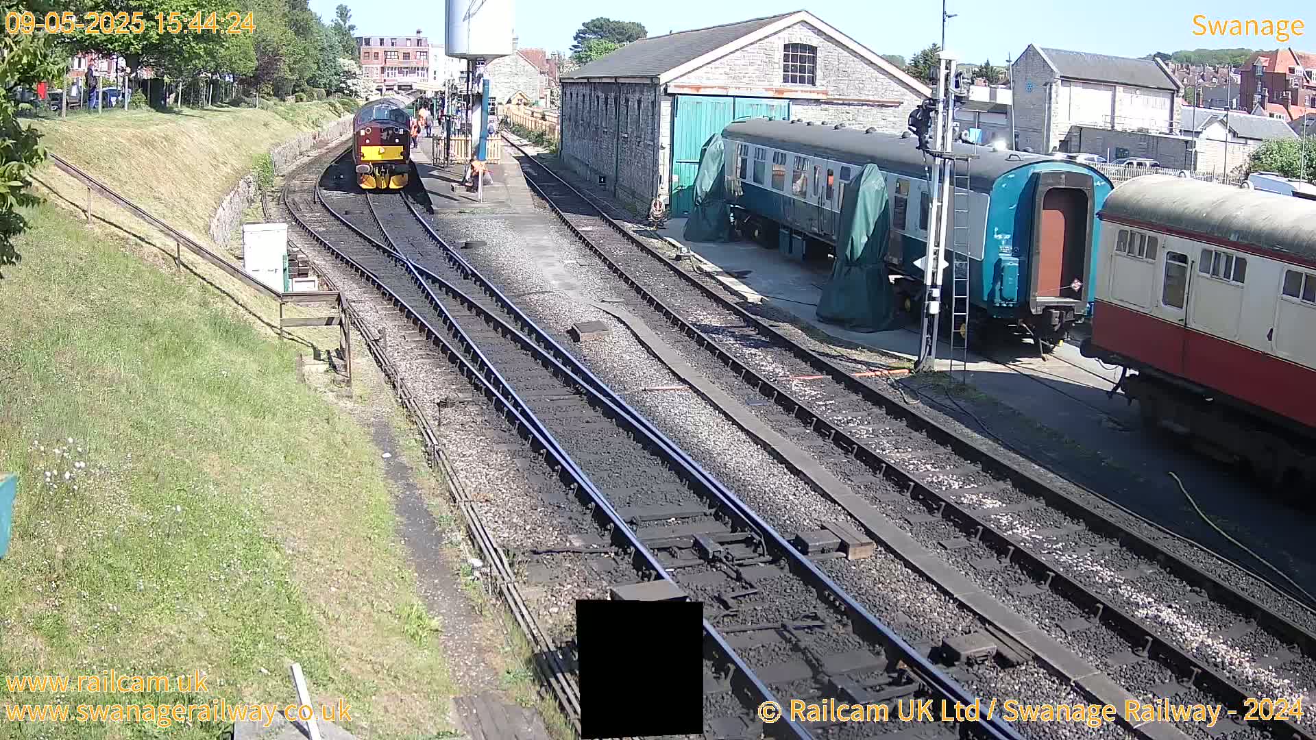 A diesel train is approaching a railway station on a sunny day, alongside several railway carriages and a stone building.