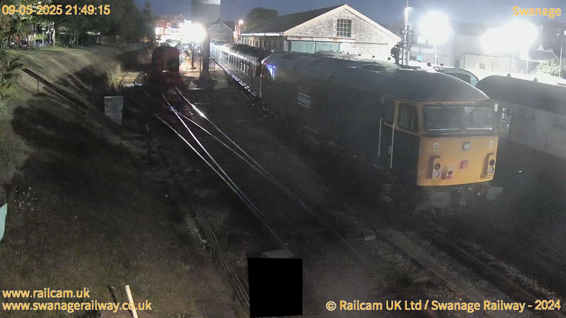 At night, a diesel locomotive and several railway carriages are parked on train tracks next to a stone building.