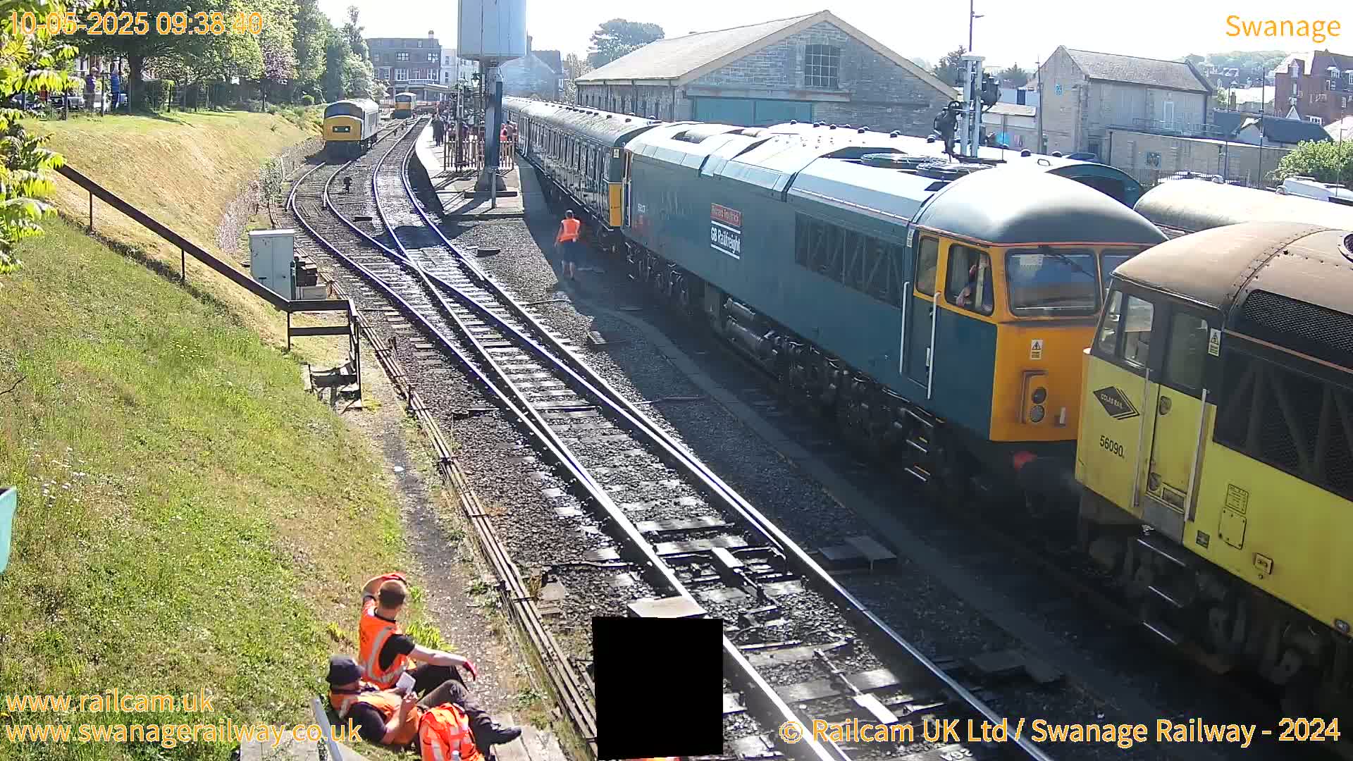 A sunny day at a train station shows several locomotives and passenger cars on multiple tracks, with some workers visible near the trains.