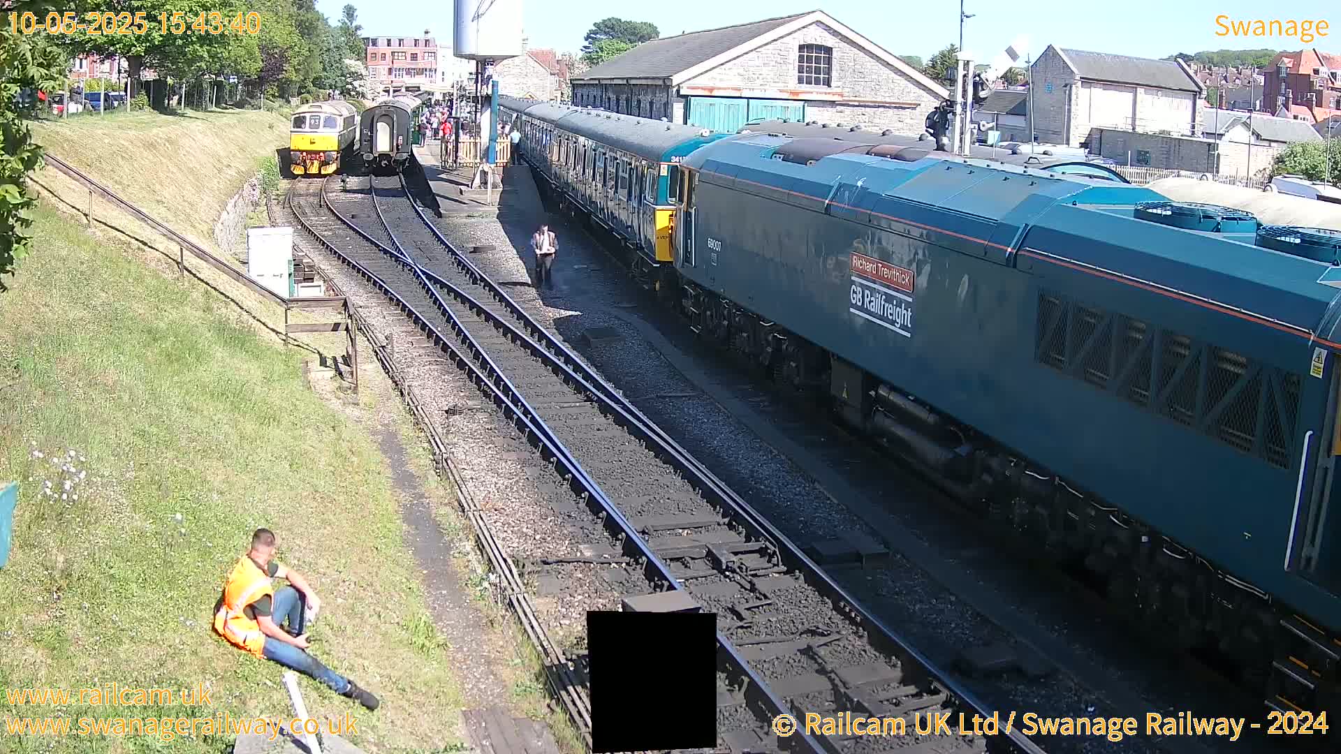 A sunny day at a train station shows two trains on parallel tracks, one a teal-colored locomotive pulling several passenger cars and the other a smaller, yellow engine with a few cars behind it.