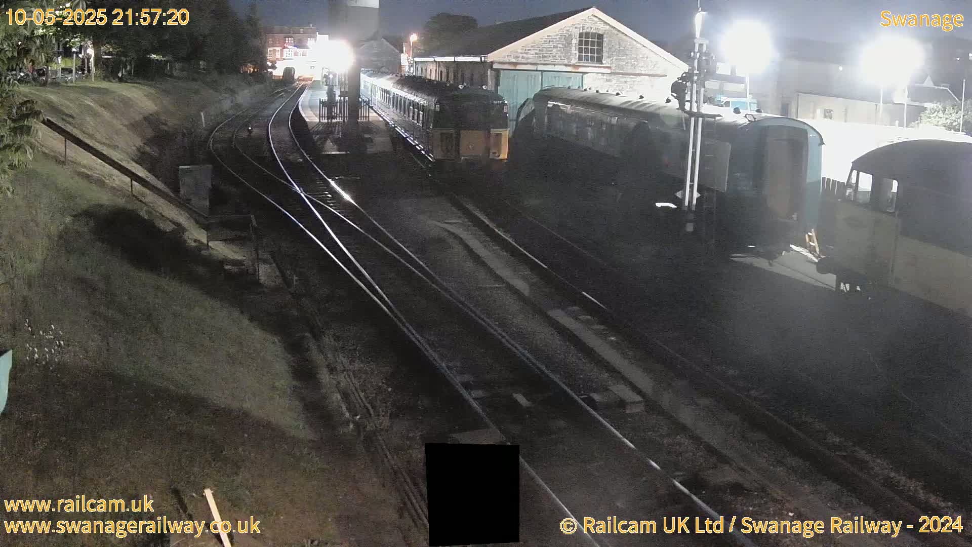 At night, several train cars are parked on railroad tracks next to a building, under artificial light.