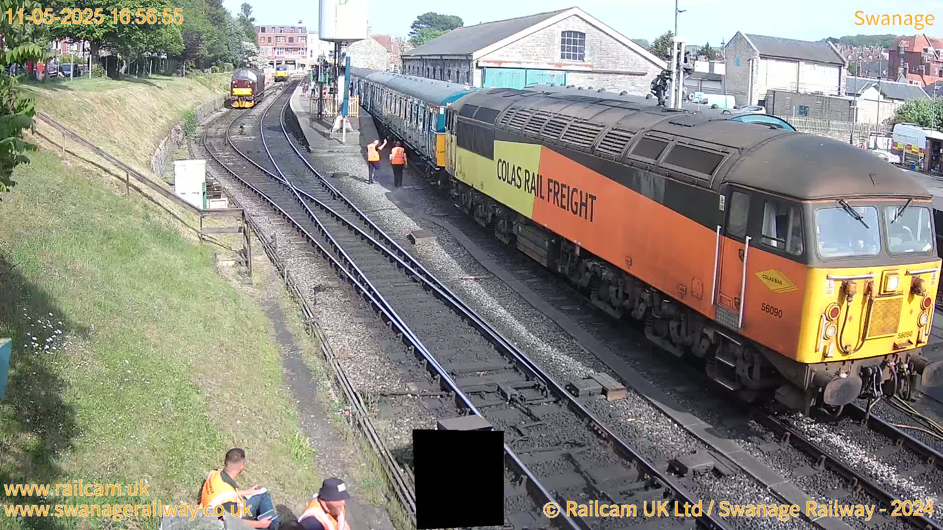 A long freight train, pulled by an orange and black locomotive, sits at a railway station on a sunny day.