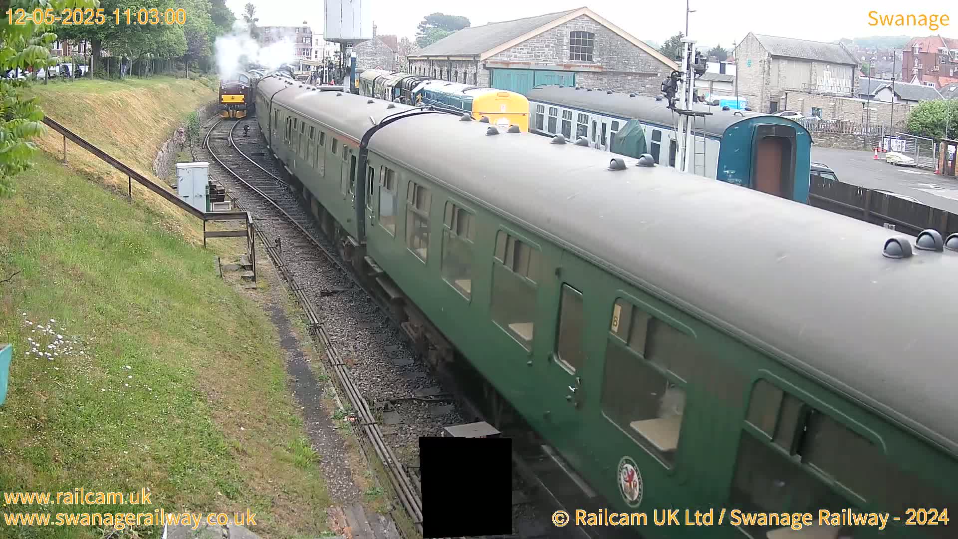 A long green passenger train is moving along a railway track, with a steam train visible in the distance on an overcast day.