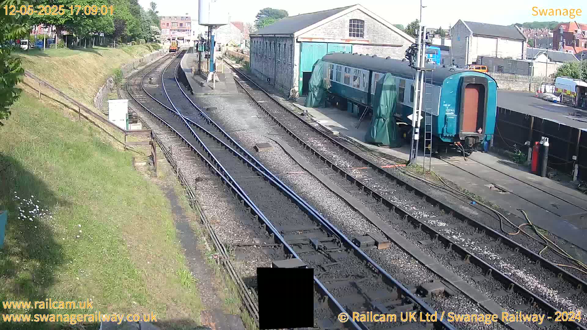 A sunny day at a railway station shows train cars parked next to a stone building and several railway tracks extending into the distance.