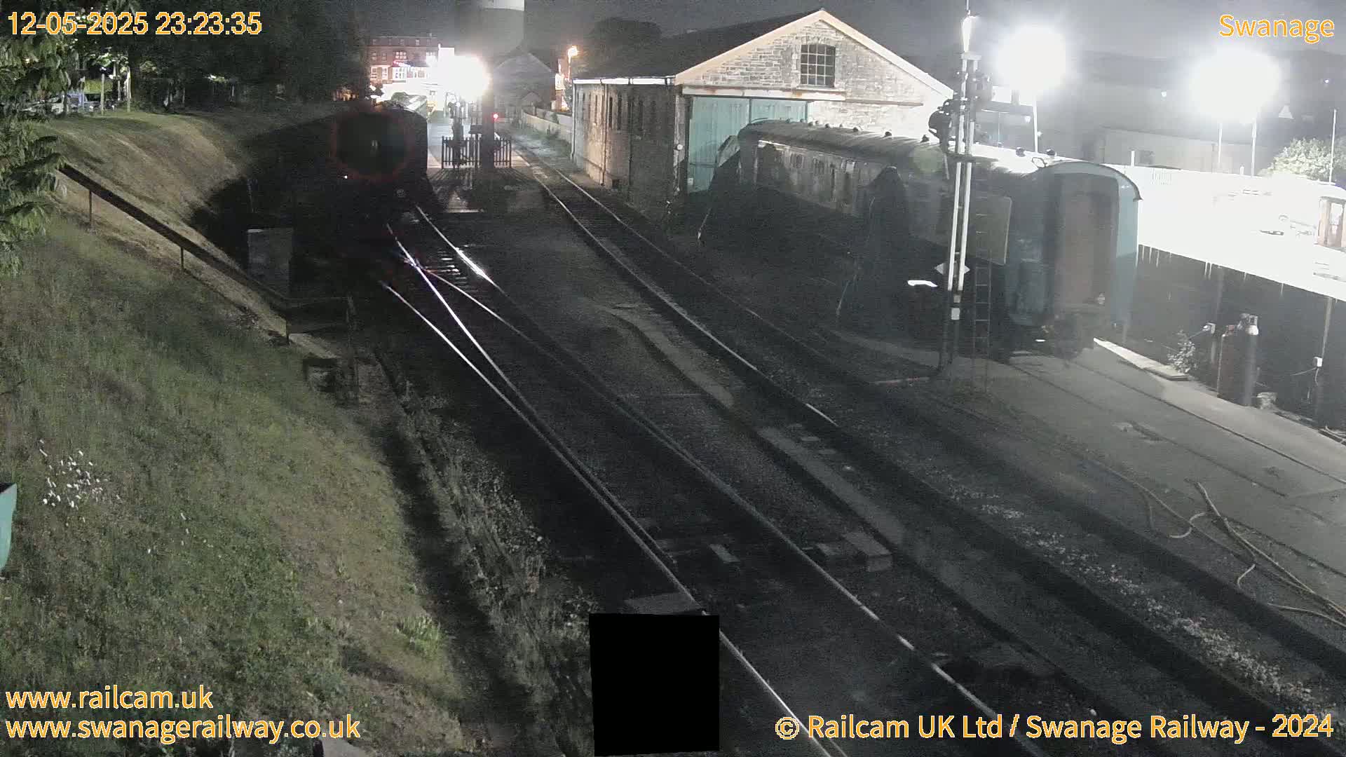 At night, a railway yard shows a train car parked next to a stone building, with train tracks extending into the distance under artificial light.