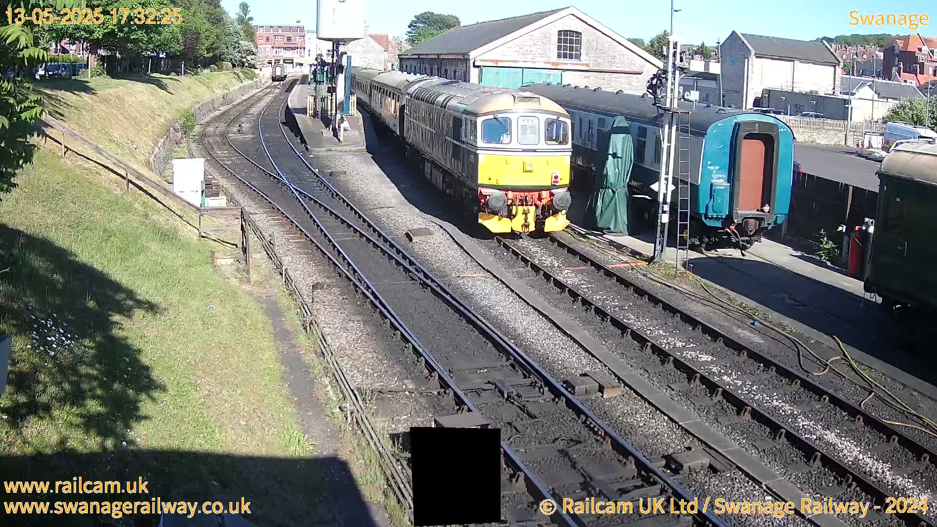 A diesel train sits at a station on a sunny day next to several other railway cars and buildings.