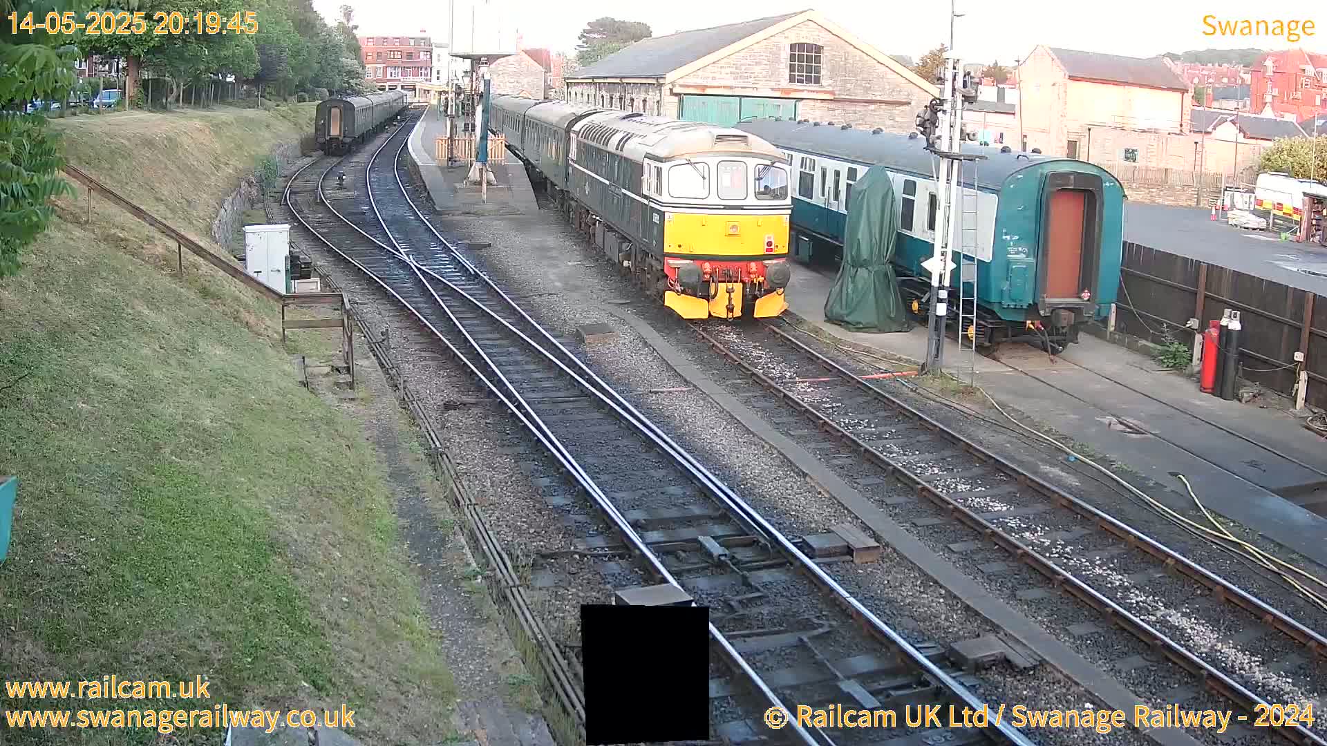 A diesel locomotive leads a train of passenger cars at a railway station on a partly cloudy evening.